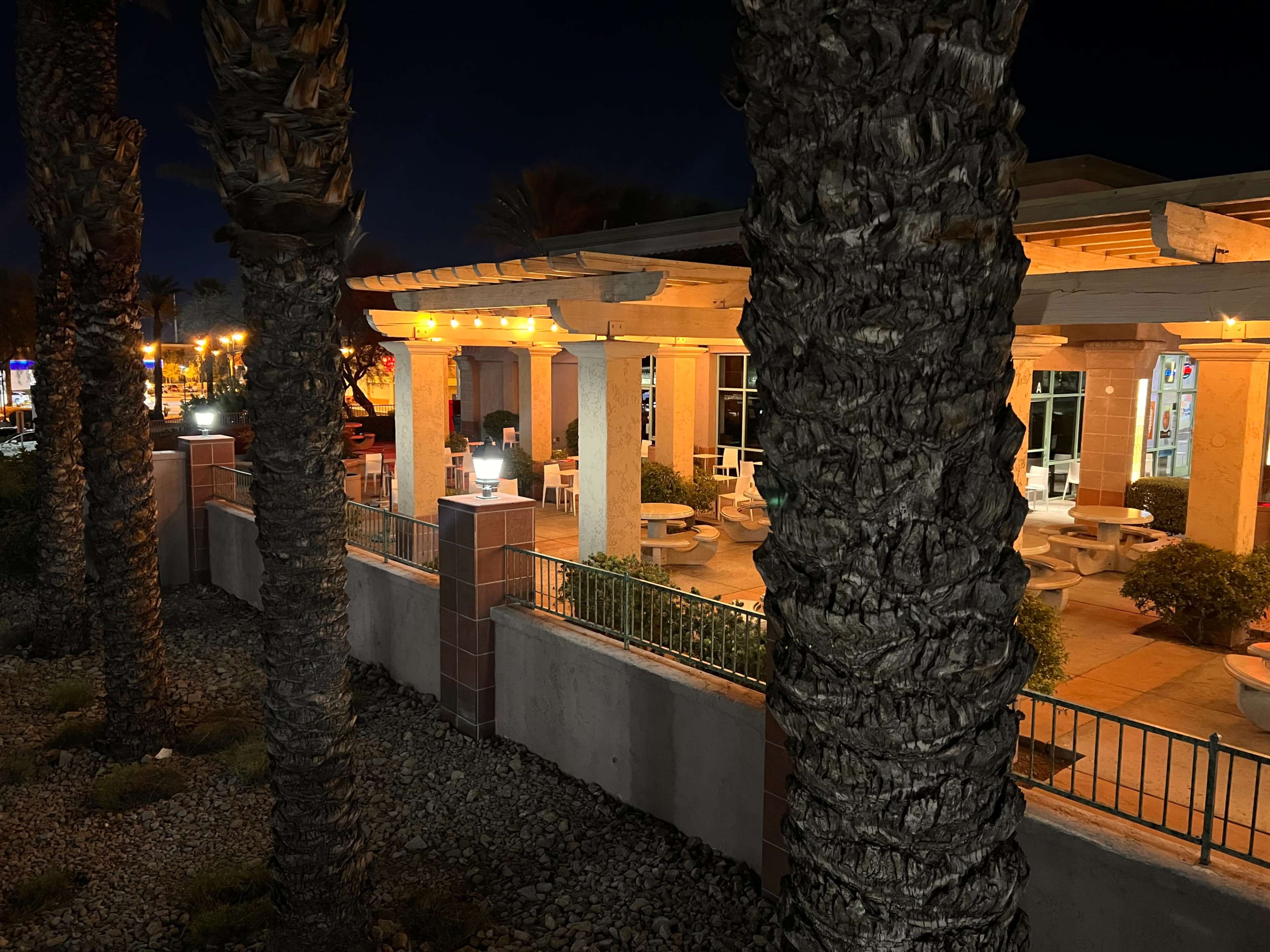 A dining establishment with outdoor seating is illuminated by string lights at night, surrounded by palm trees and rocky landscaping.