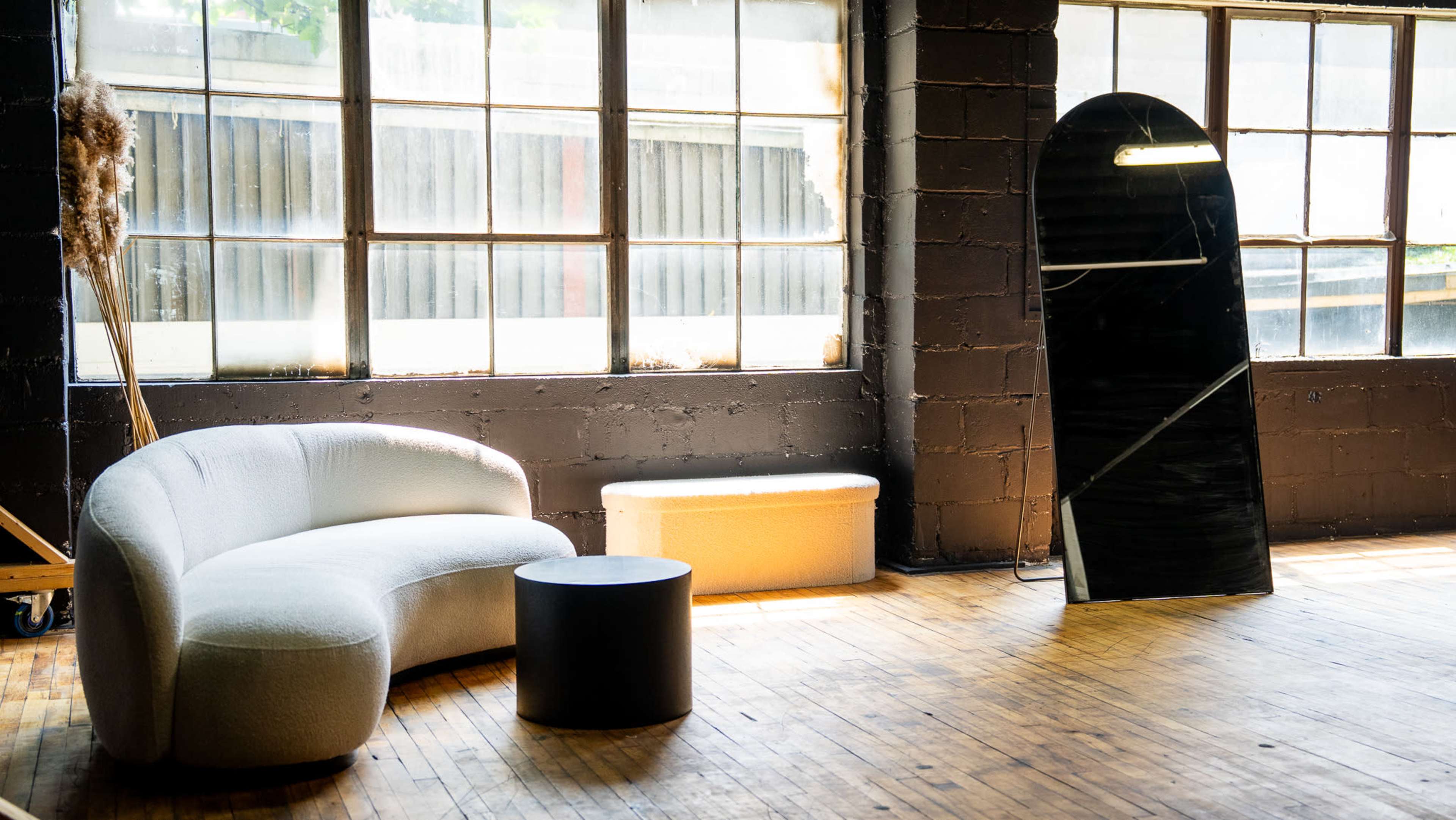 The image features a modern, minimalist interior with a curved sofa, a round coffee table, and a large, reflective mirror positioned near tall windows that allow natural light to enter.