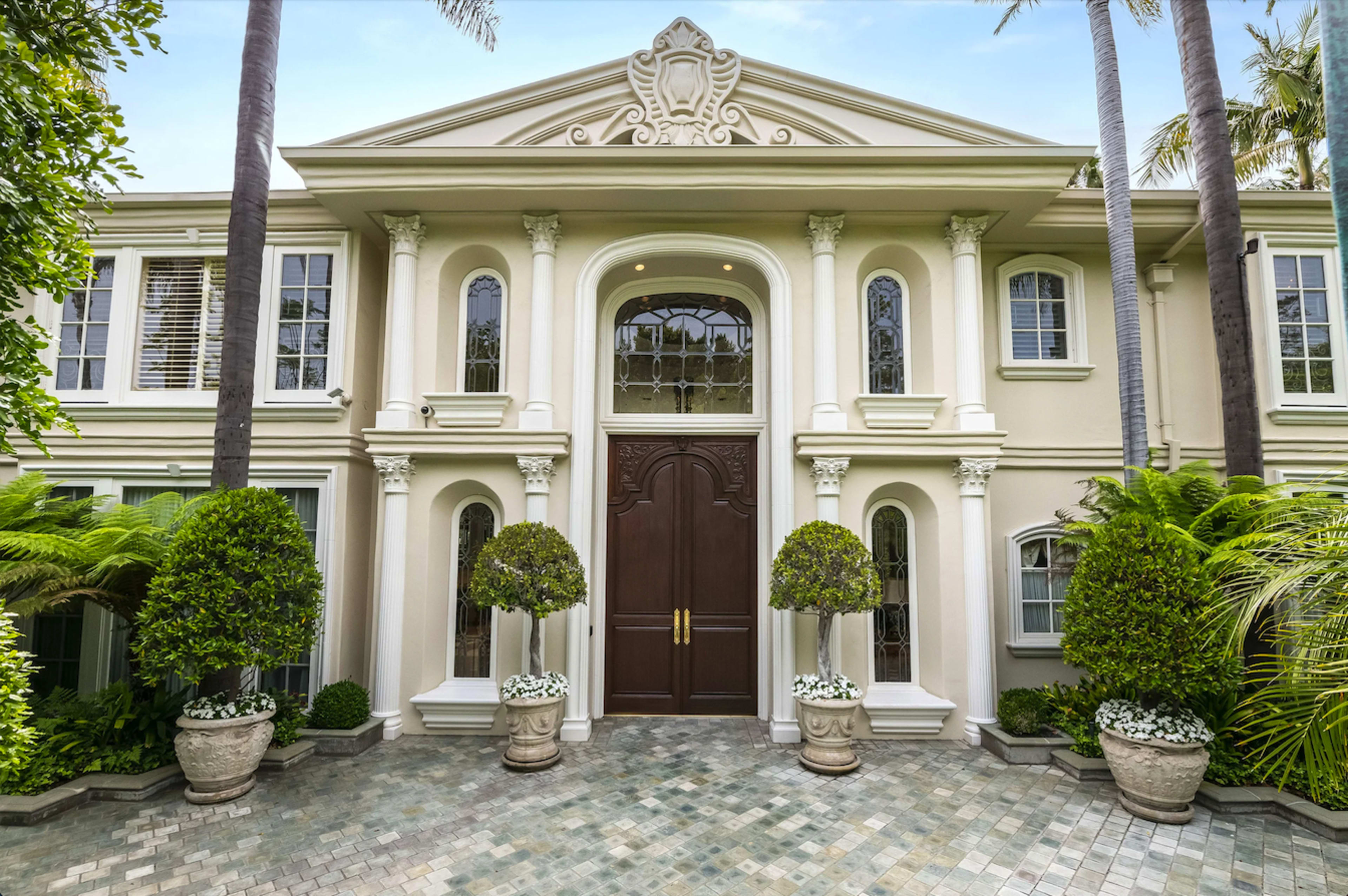 The image shows a grand entrance of a modern mansion featuring ornate architectural details, large double doors, and neatly landscaped greenery on either side.