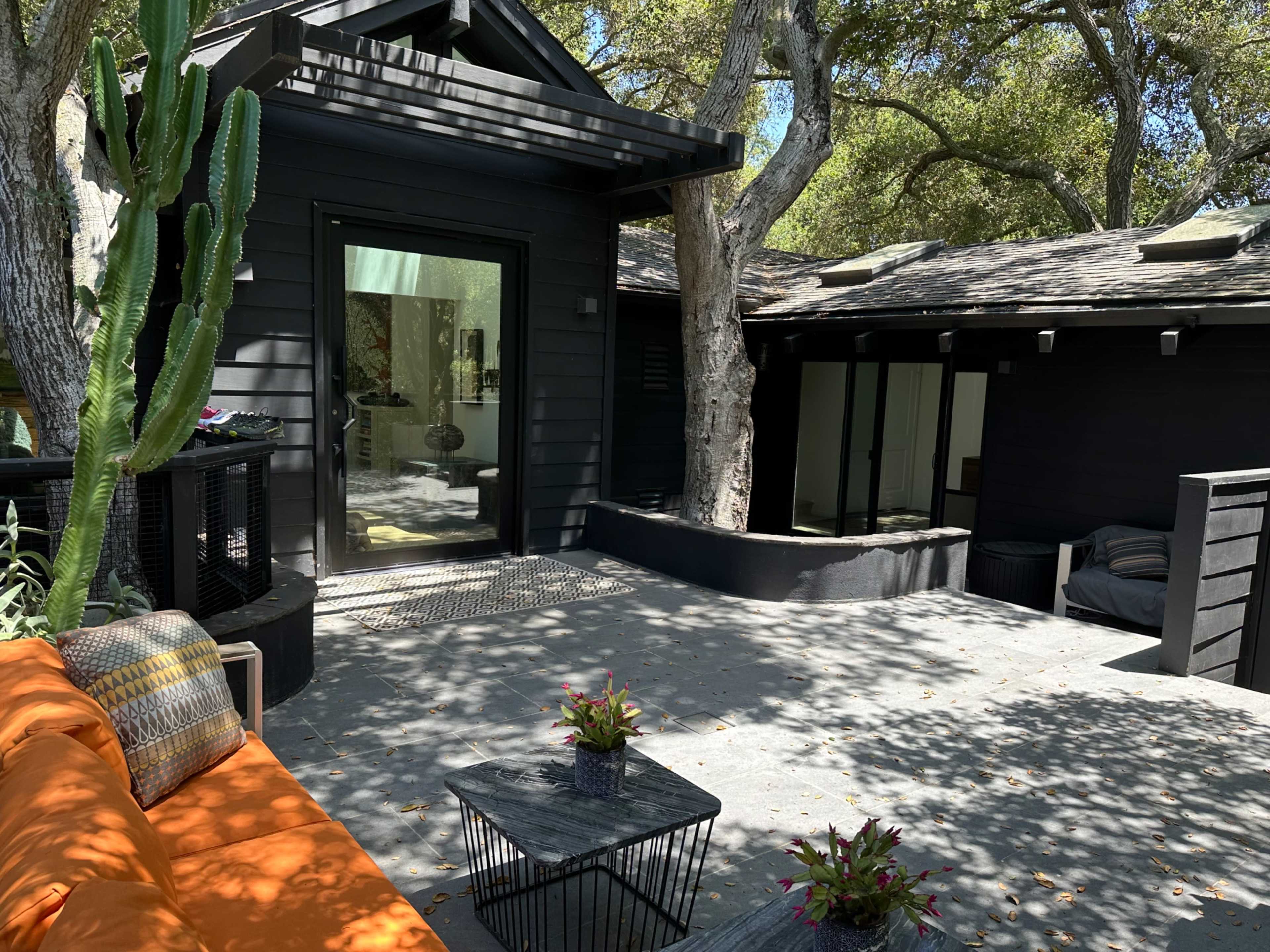 The image shows a modern black exterior house surrounded by trees, featuring a patio with orange seating and potted plants.