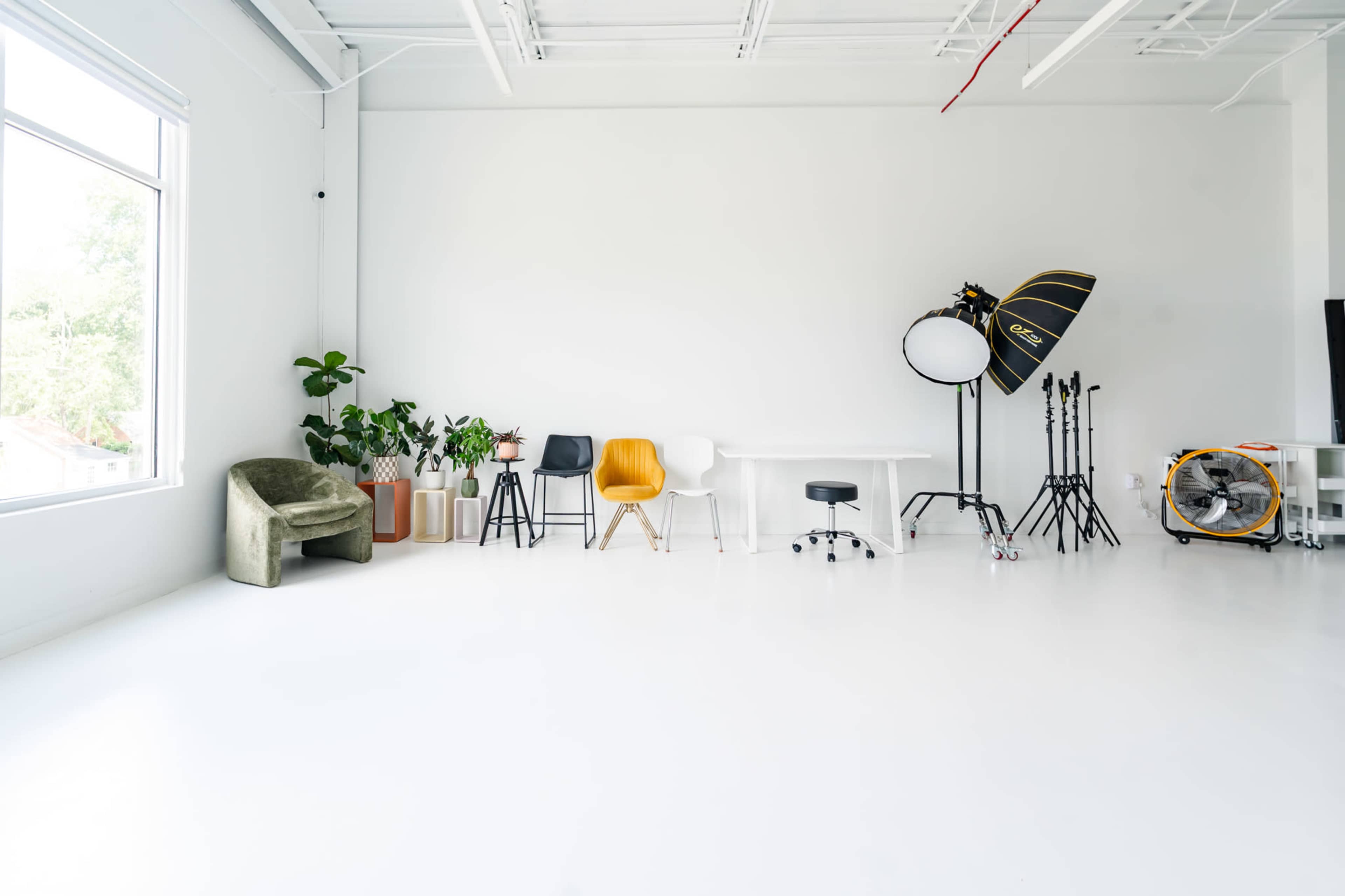 The image features a spacious, well-lit studio with white walls and a polished floor, furnished with various chairs, a table, and photography equipment.