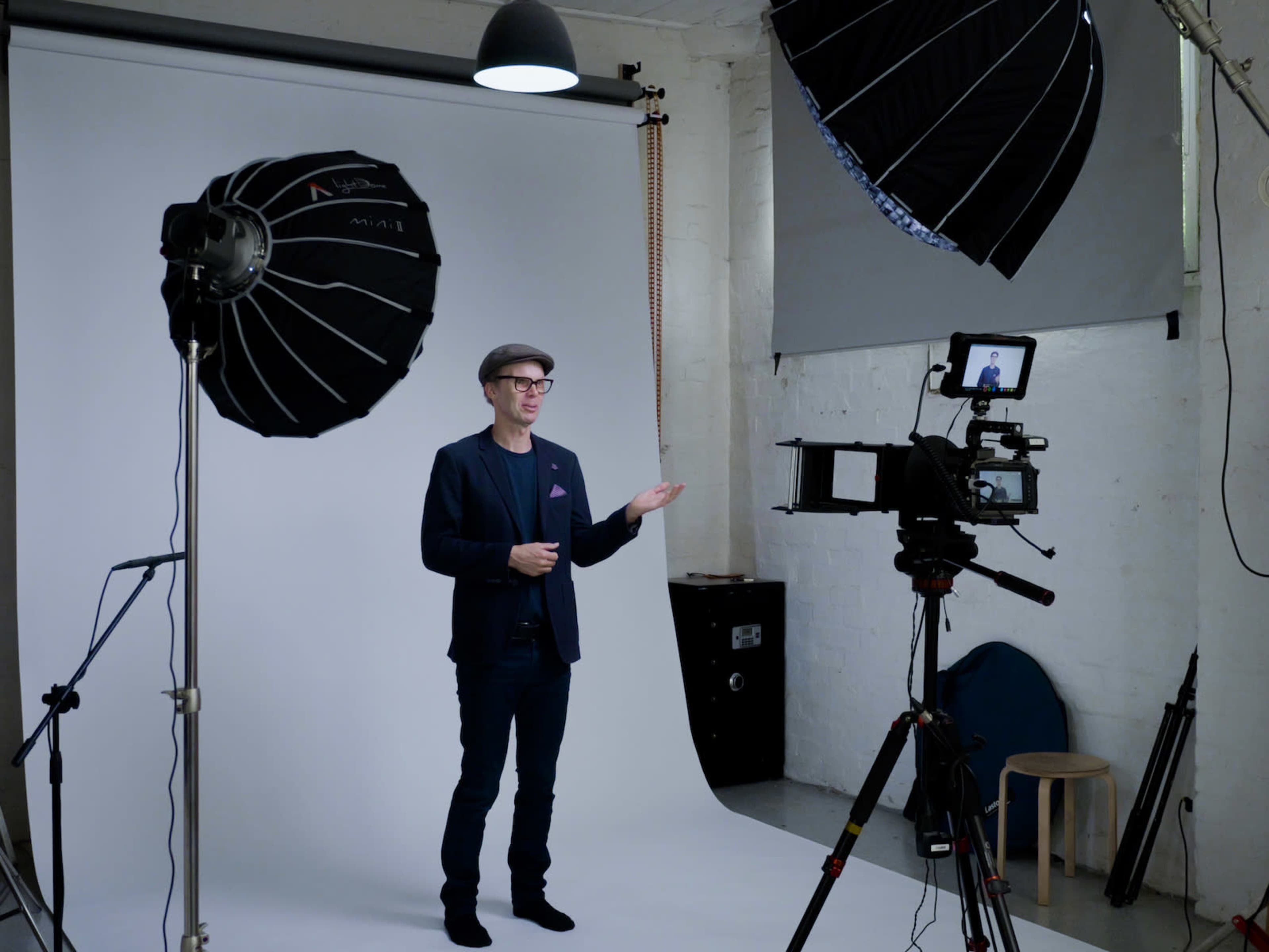 A man stands in front of a backdrop in a photography studio, gesturing towards a camera mounted on a tripod.