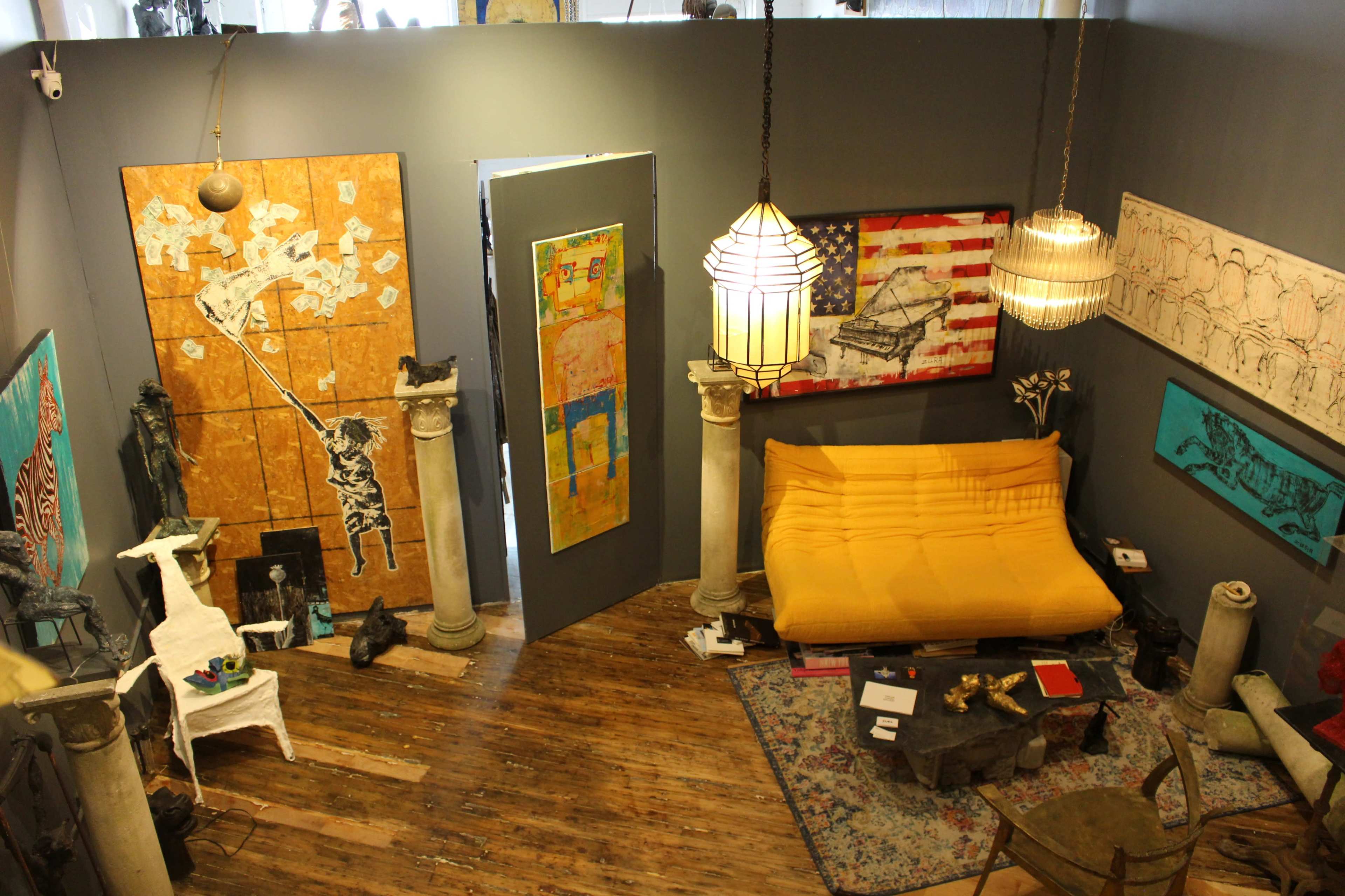 The image shows an eclectic art gallery featuring a yellow sofa, various artworks on the walls, and distinctive lighting fixtures.
