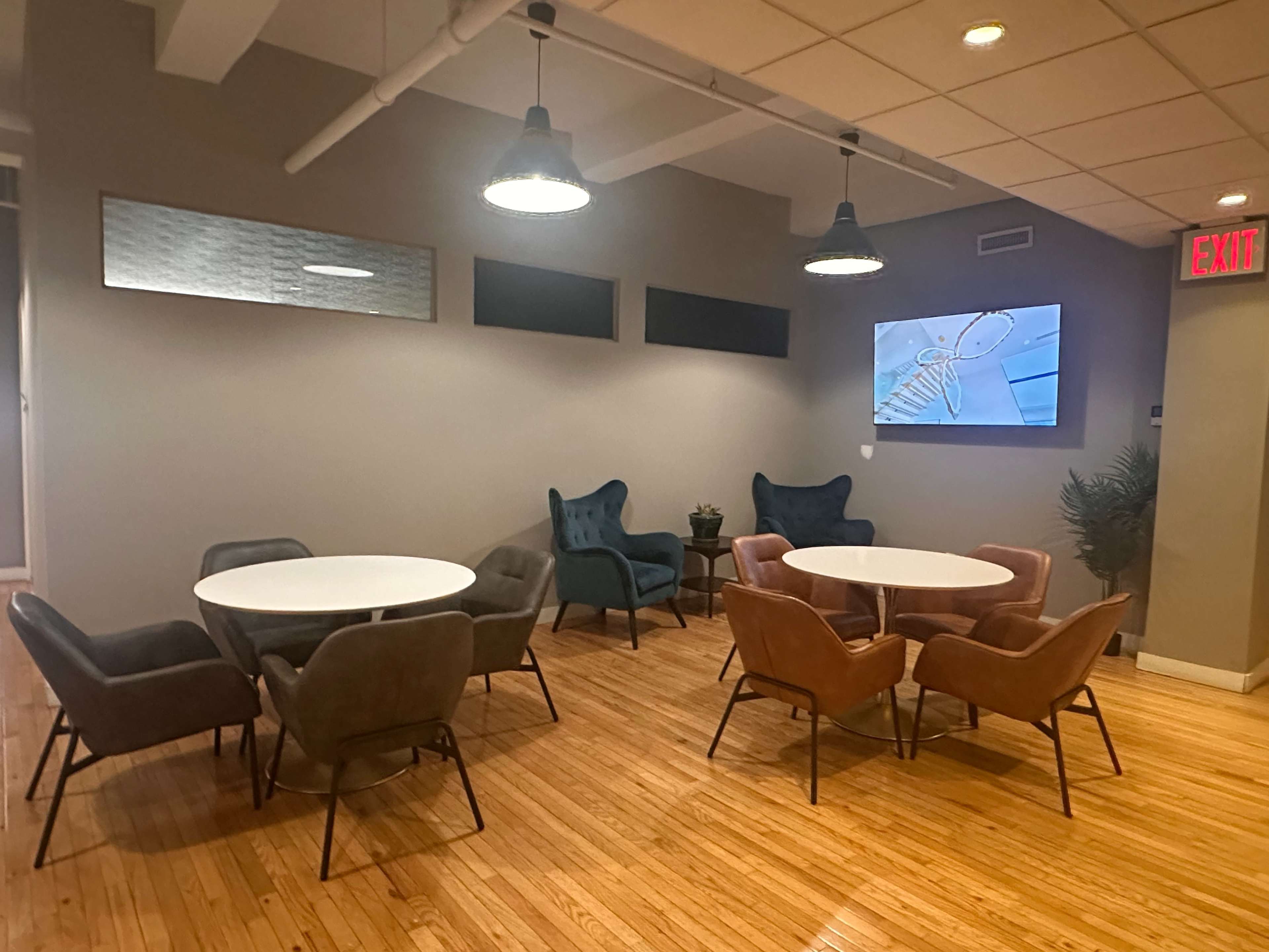 The image shows a modern lounge area with multiple round tables surrounded by chairs in a space with wooden flooring and a wall-mounted television displaying an animated character.