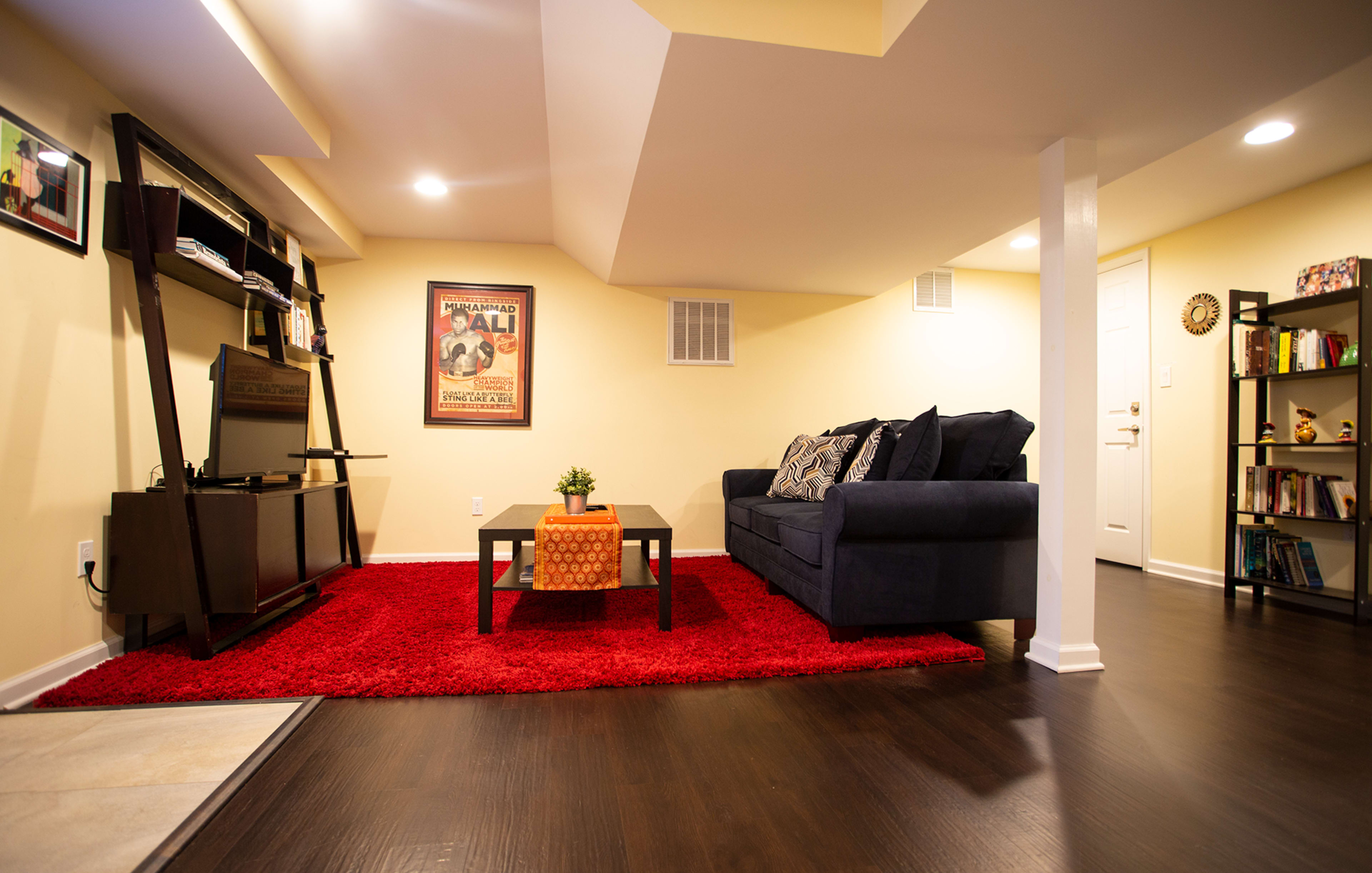 The image shows a neatly arranged basement living area with a dark blue sofa, a red area rug, a small coffee table, a television stand, and shelves filled with books.