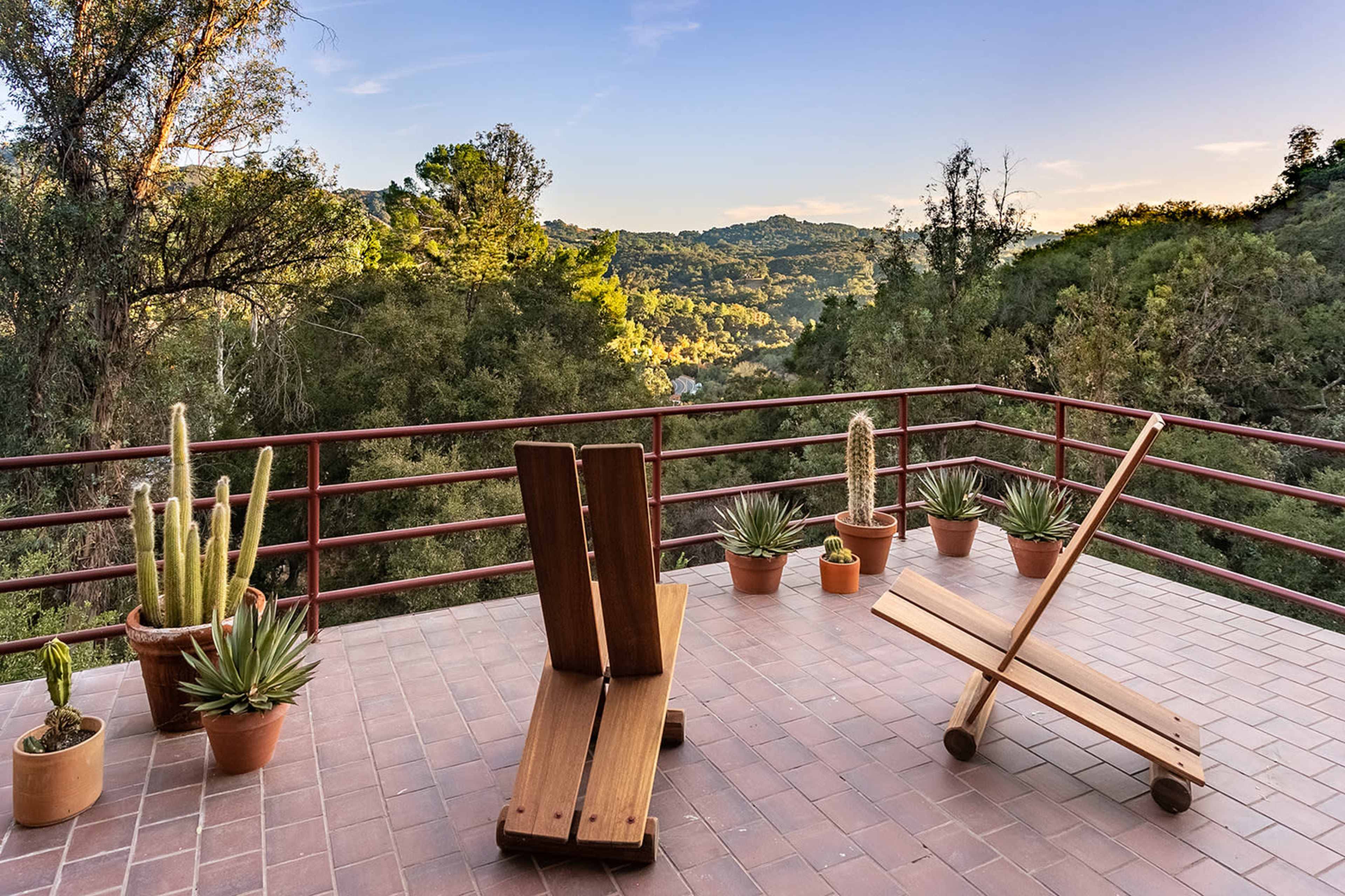 A wooden terrace features two lounge chairs surrounded by potted plants and a scenic view of a green landscape.