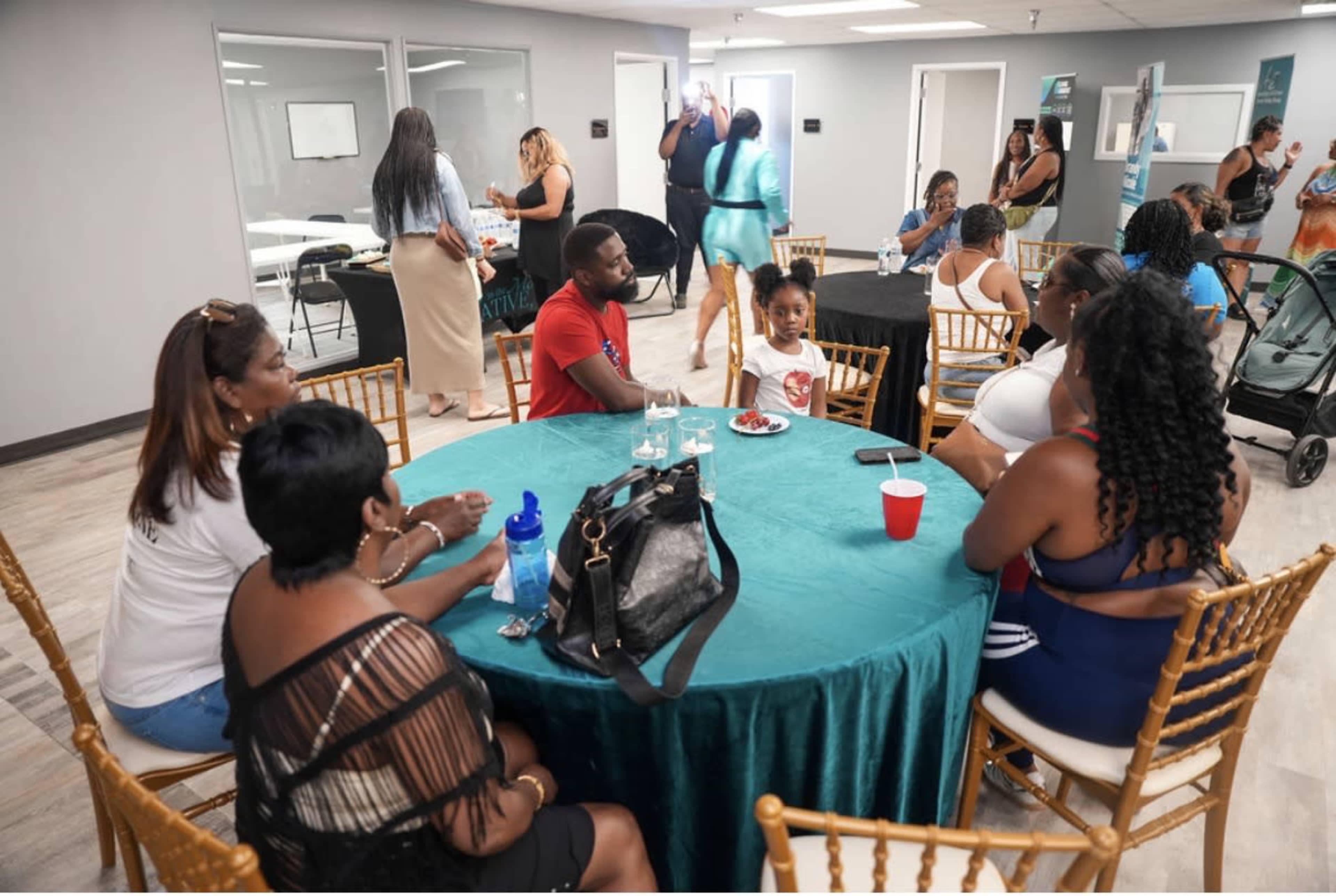 The image shows a gathering in a brightly lit room with several people seated around tables, while a child plays in a costume in the background.