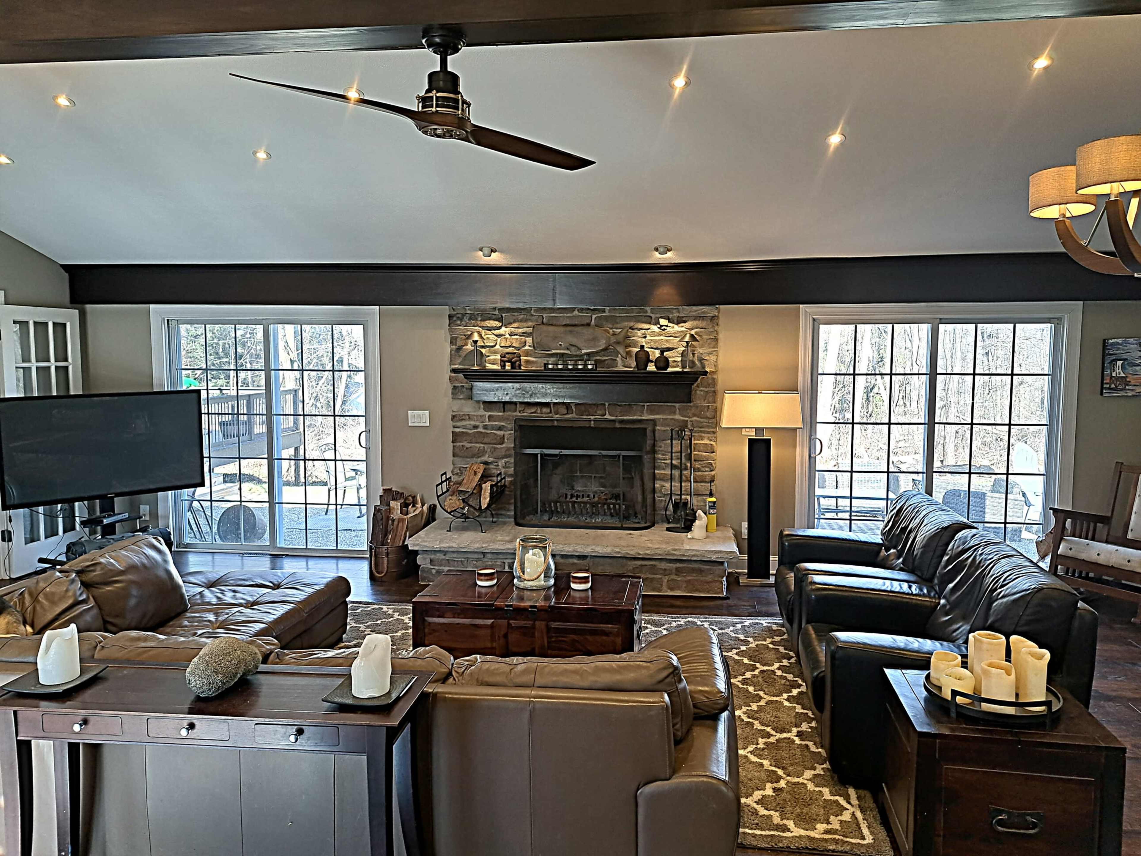 A cozy living room features a stone fireplace, two brown leather sofas, a wooden coffee table, and large windows that provide natural light.