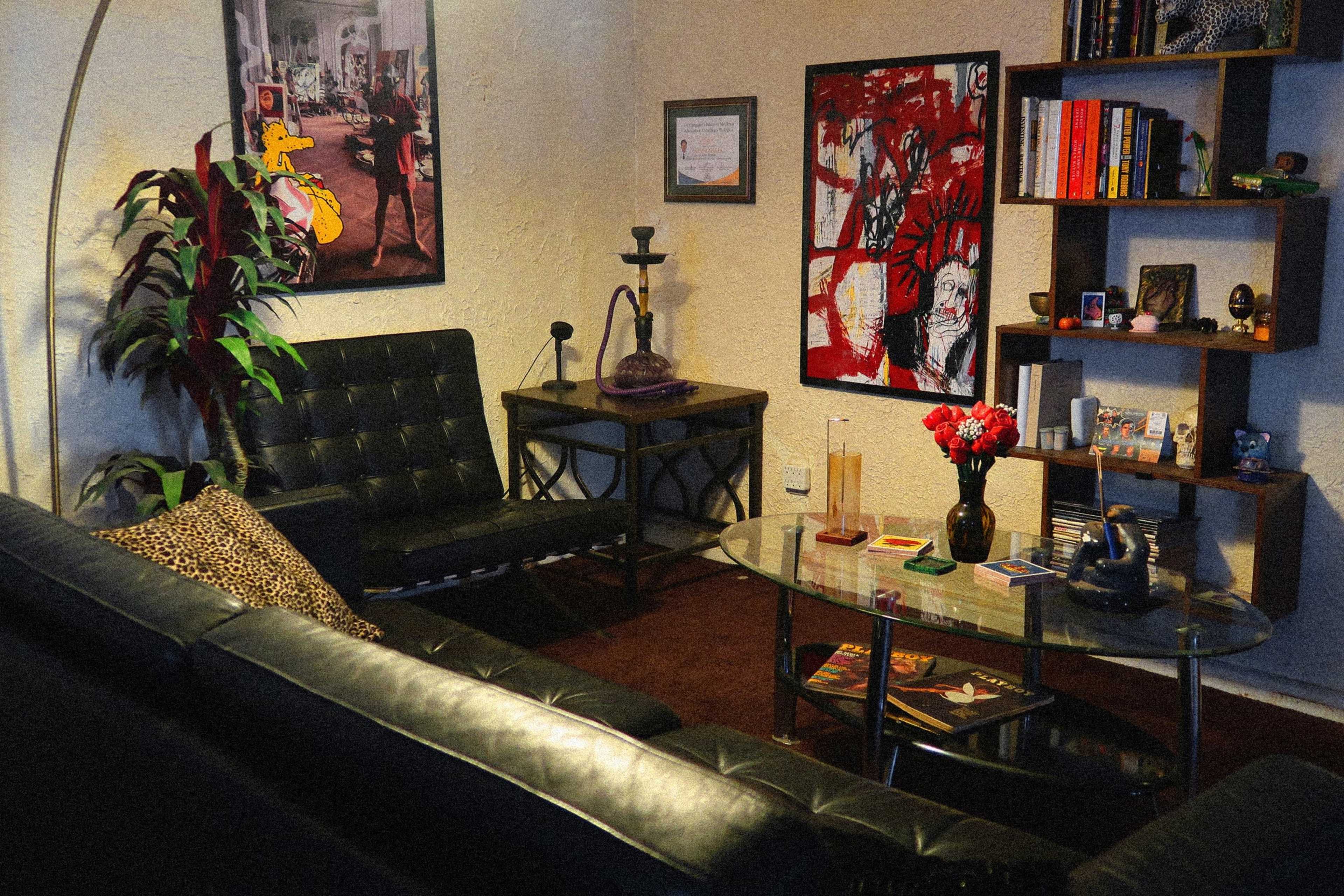The image depicts a cozy living room with a black leather sofa, a glass coffee table, colorful artwork on the walls, and a small shelf filled with books and decorative items.