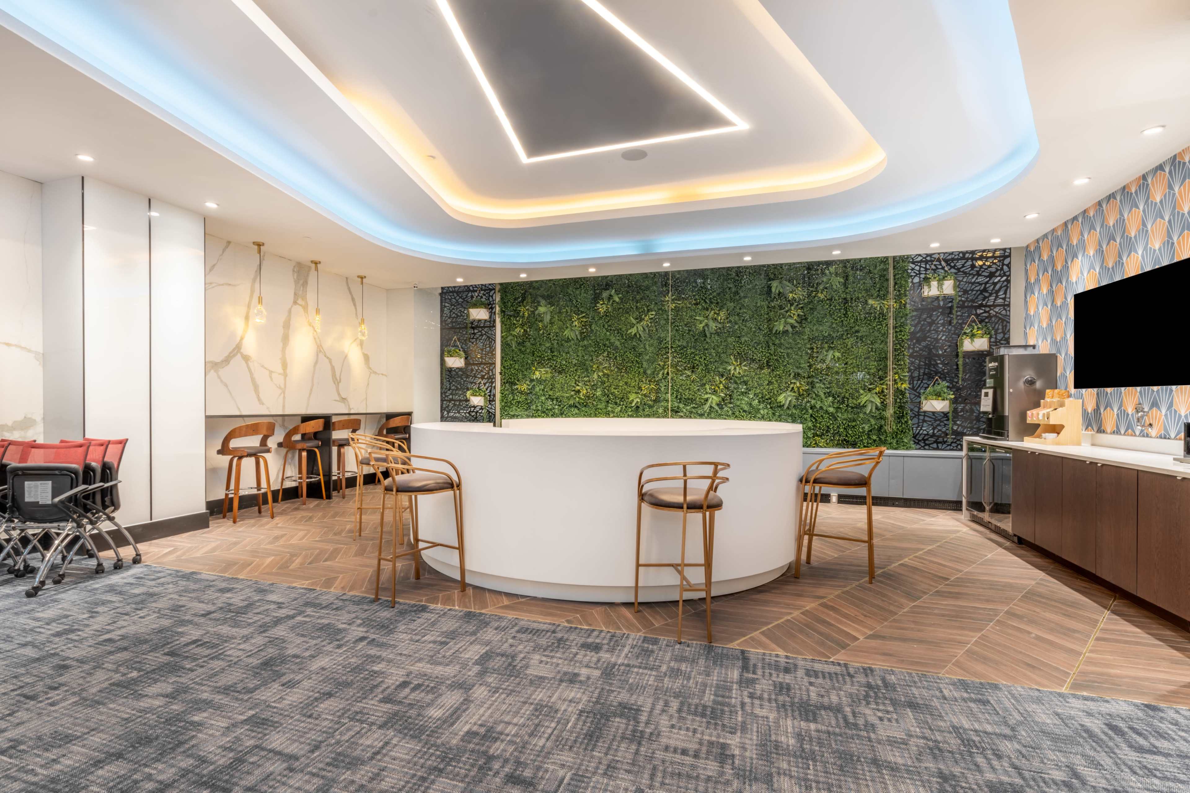 The image shows a modern lounge area featuring a curved white bar, green wall plants, wooden furniture, and a stylish ceiling design.