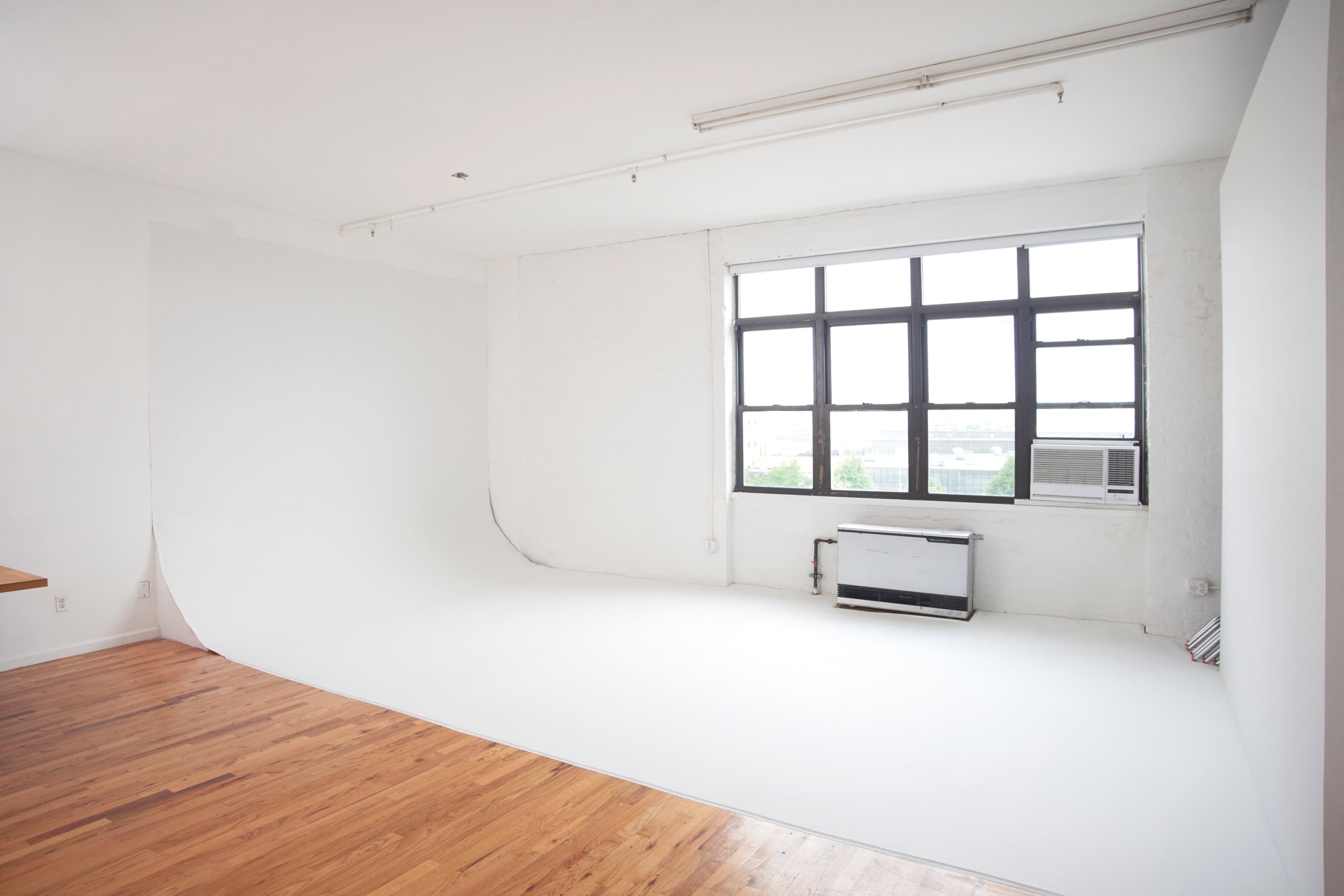 The image shows a spacious, empty room with wooden floors and a large window, featuring a seamless white backdrop along one wall.