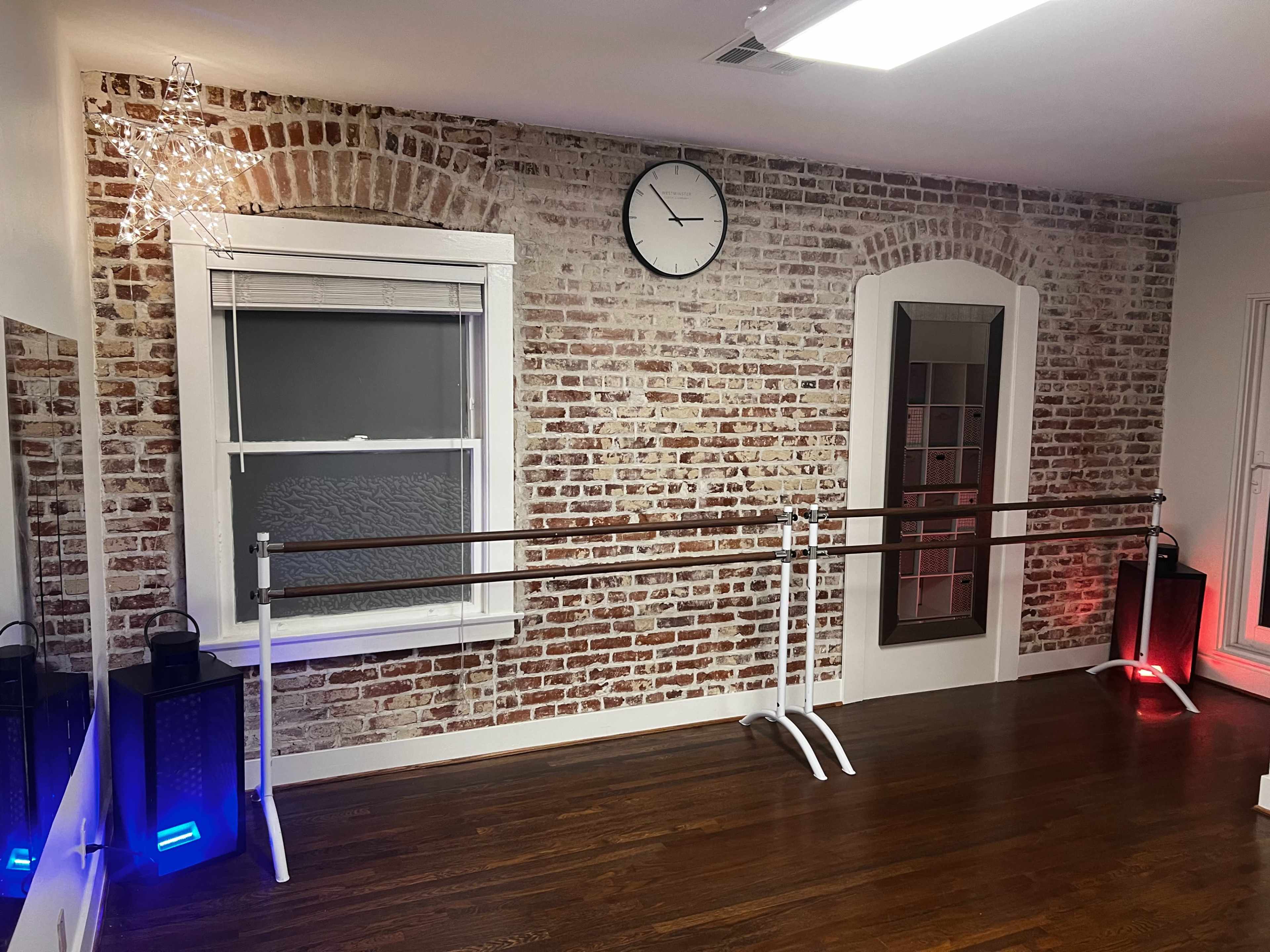 A dance studio with exposed brick walls, wooden floors, ballet barre setups, a clock on the wall, and ambient lighting.