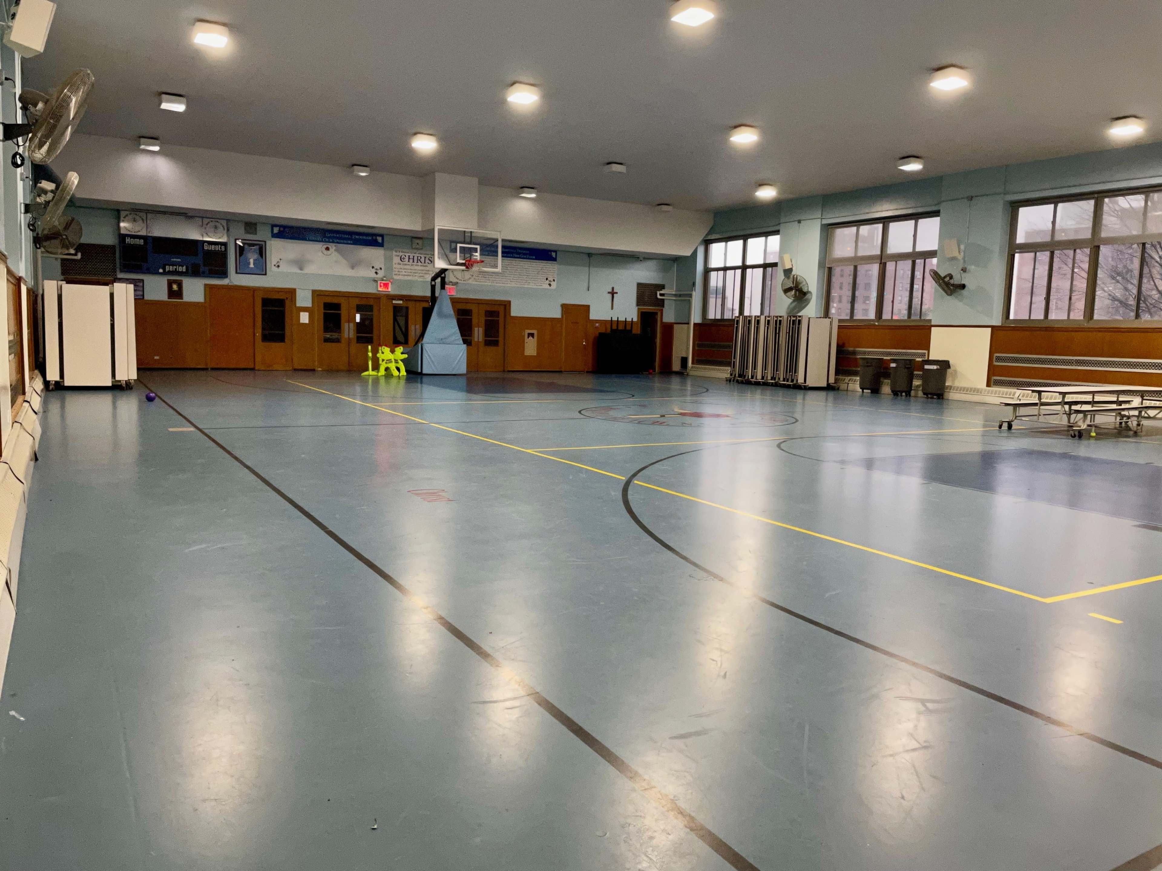 An empty school gymnasium with polished blue floors, basketball hoops, and folding tables along the side.