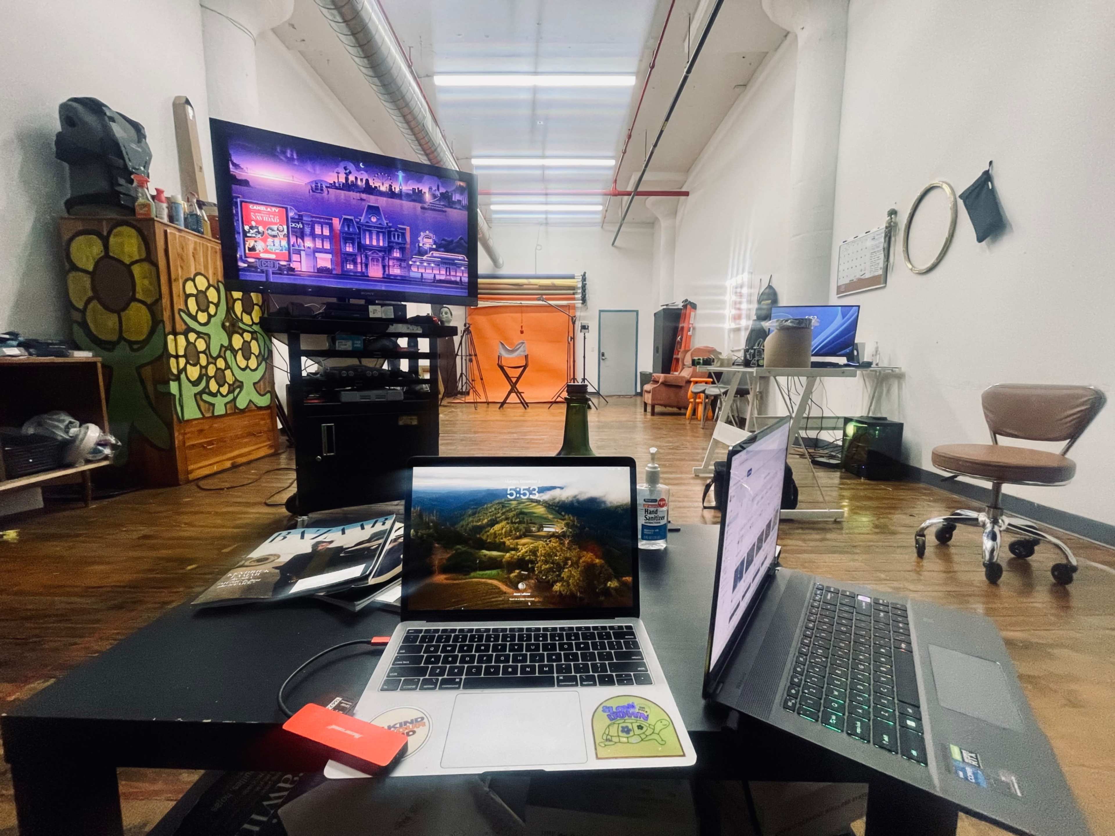 The image shows an open workspace featuring two laptops on a table, with a large TV and various equipment set up in the background.
