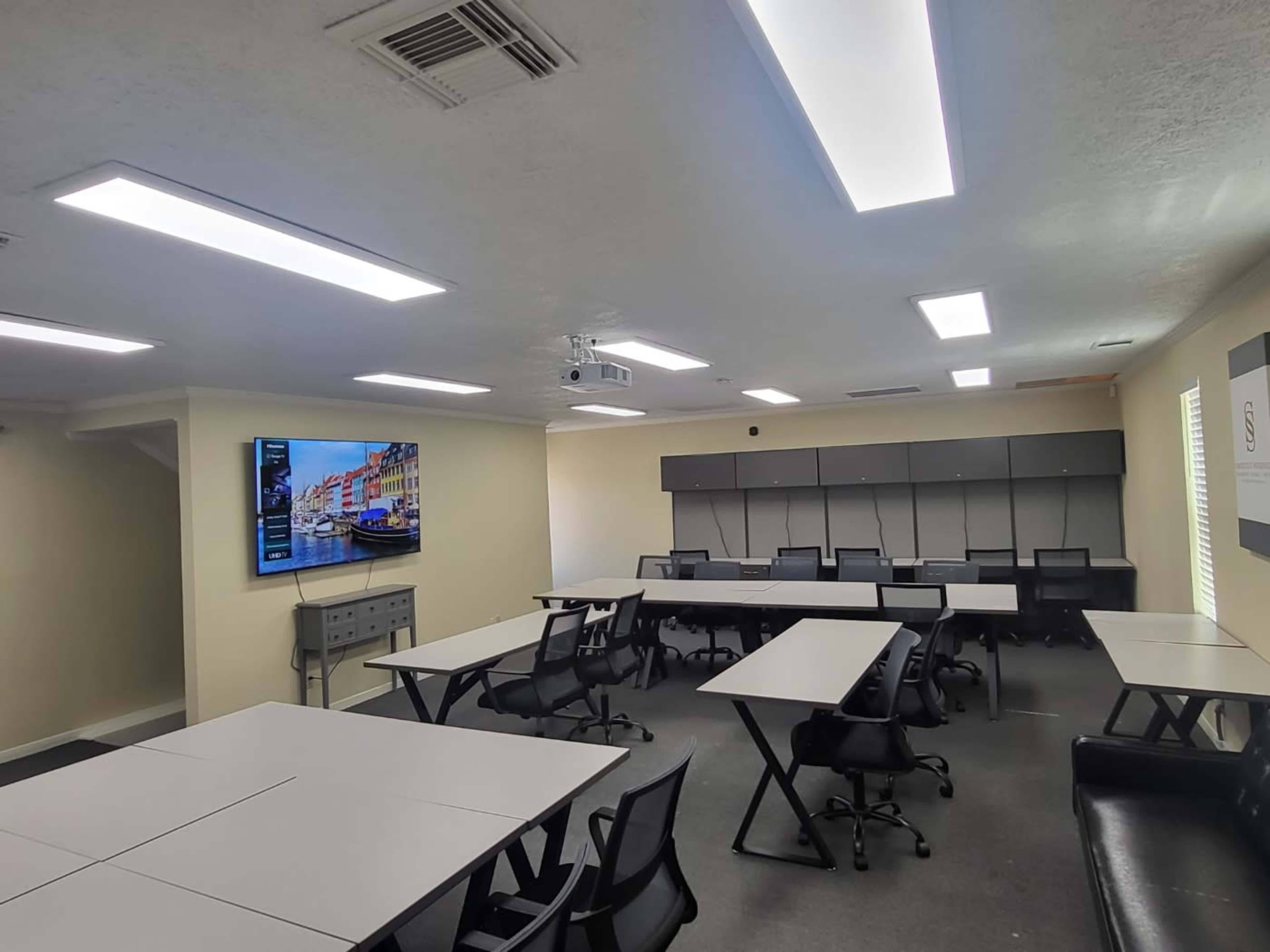 A modern meeting room with several rectangular tables, chairs, and a large wall-mounted screen displaying a cityscape.