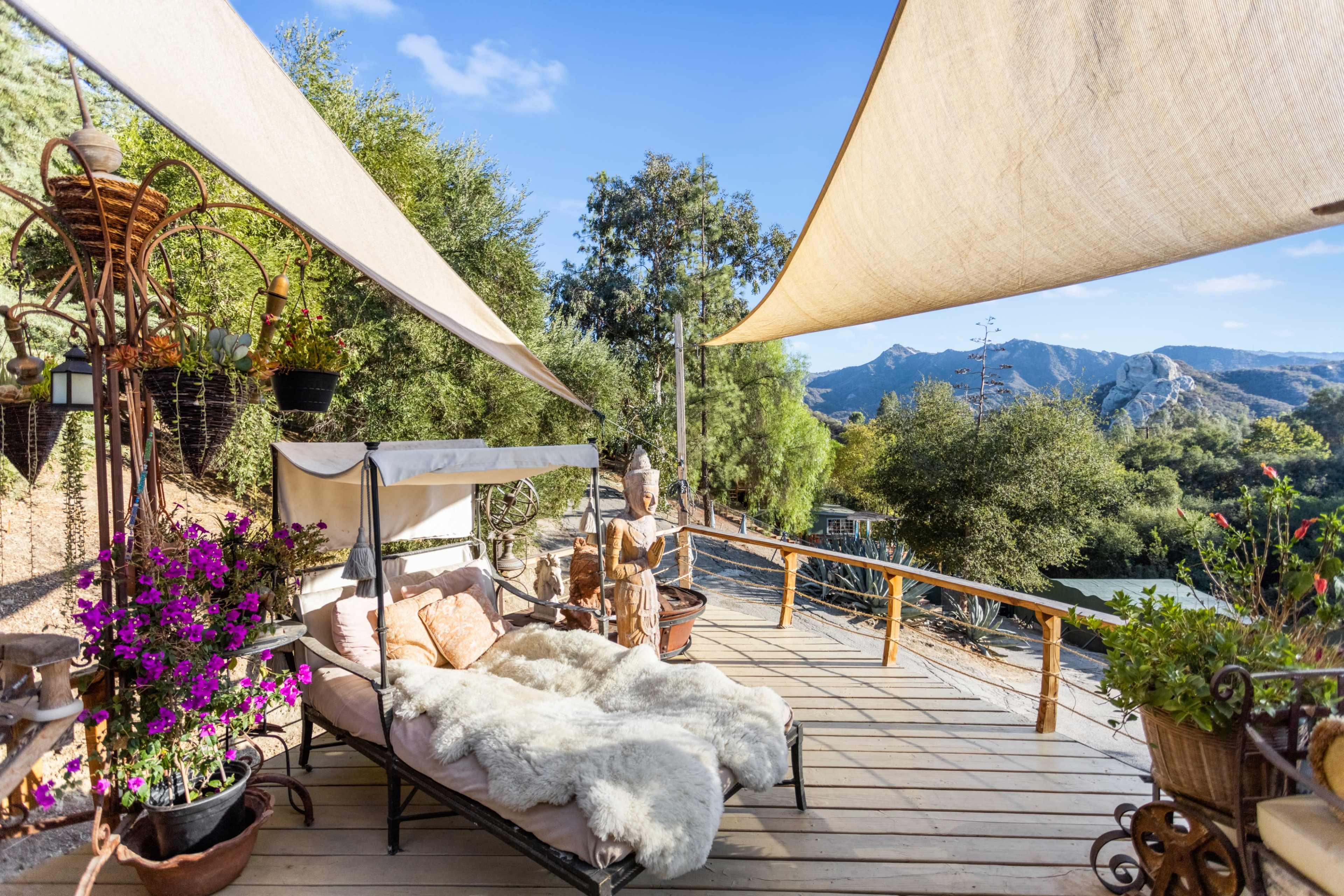 A wooden deck features a lounging area with a bed covered in a fur blanket, surrounded by potted plants and a statue, with mountains visible in the background.