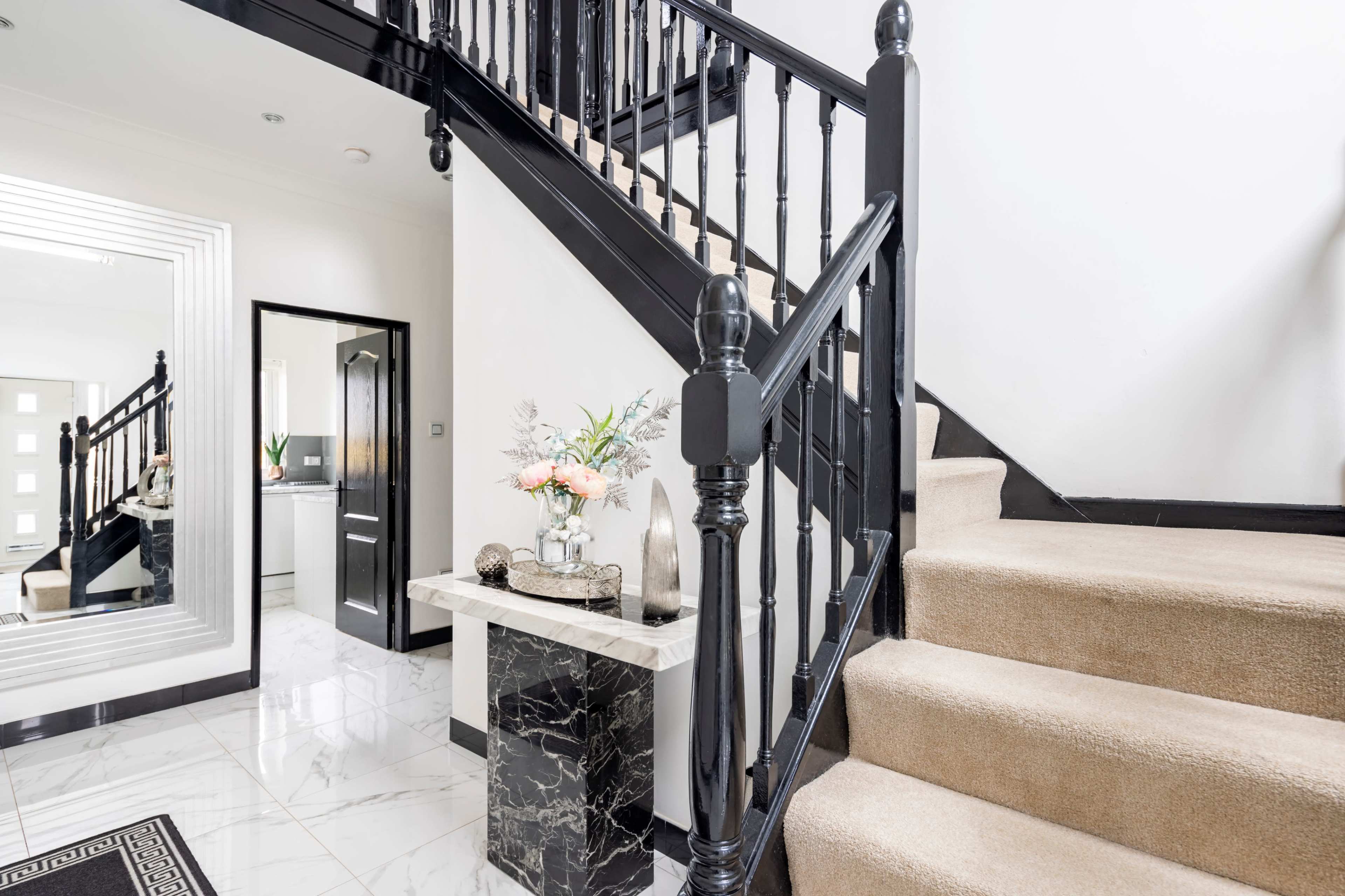 The image shows a modern staircase with black railings and a beige carpet, leading upward beside a marble-topped table in a well-lit hallway.