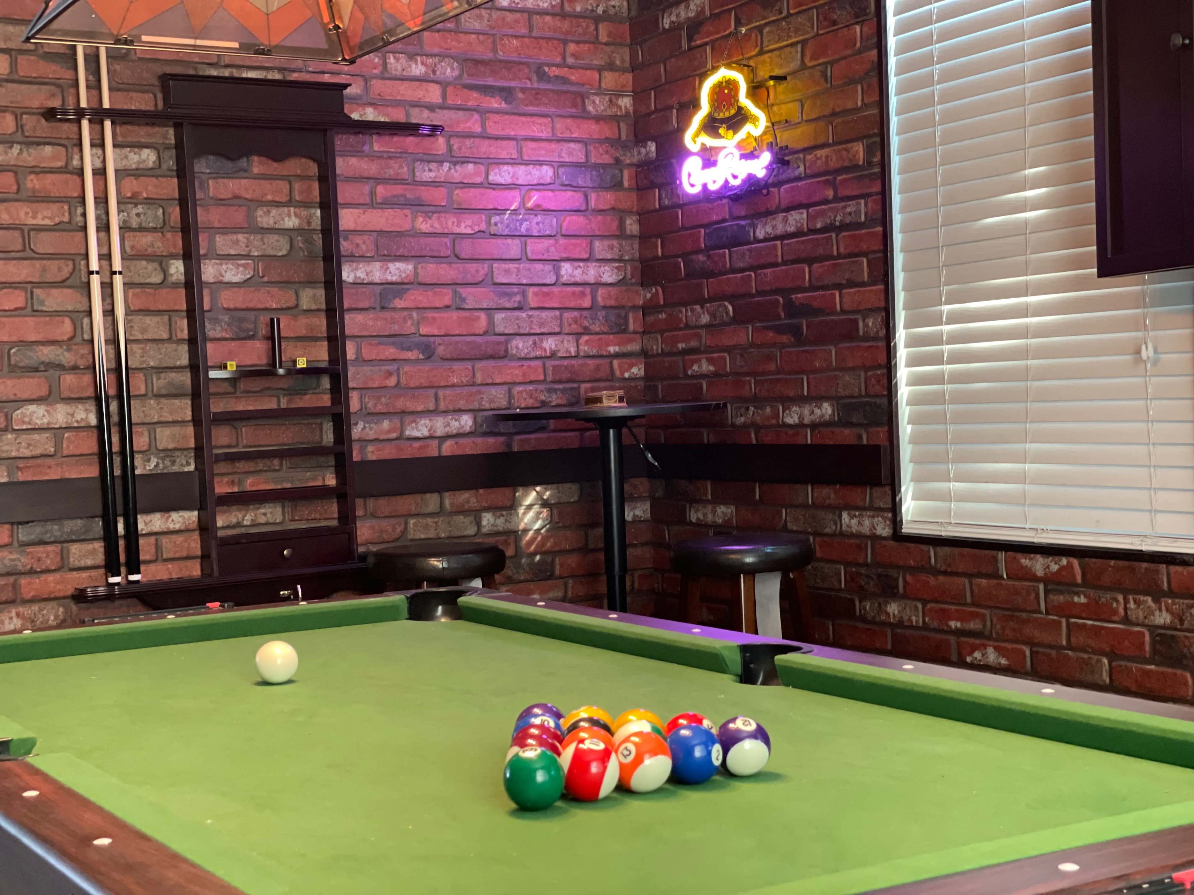 A pool table with colorful balls arranged in a triangle sits in a room with brick walls and a neon sign.