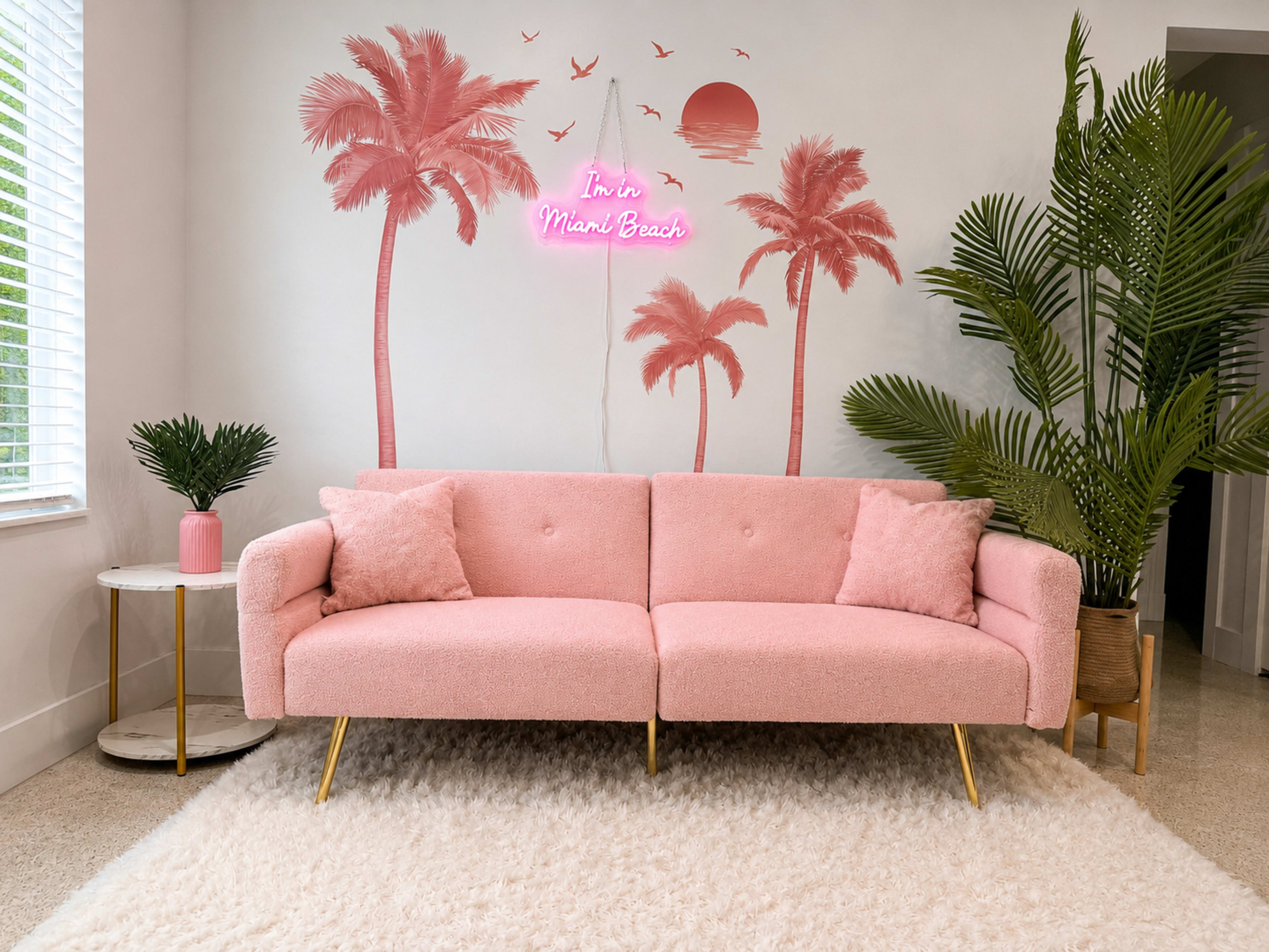 A pink sofa sits in a bright room with palm tree artwork on the wall and a neon sign that reads "I'm in Miami Beach."