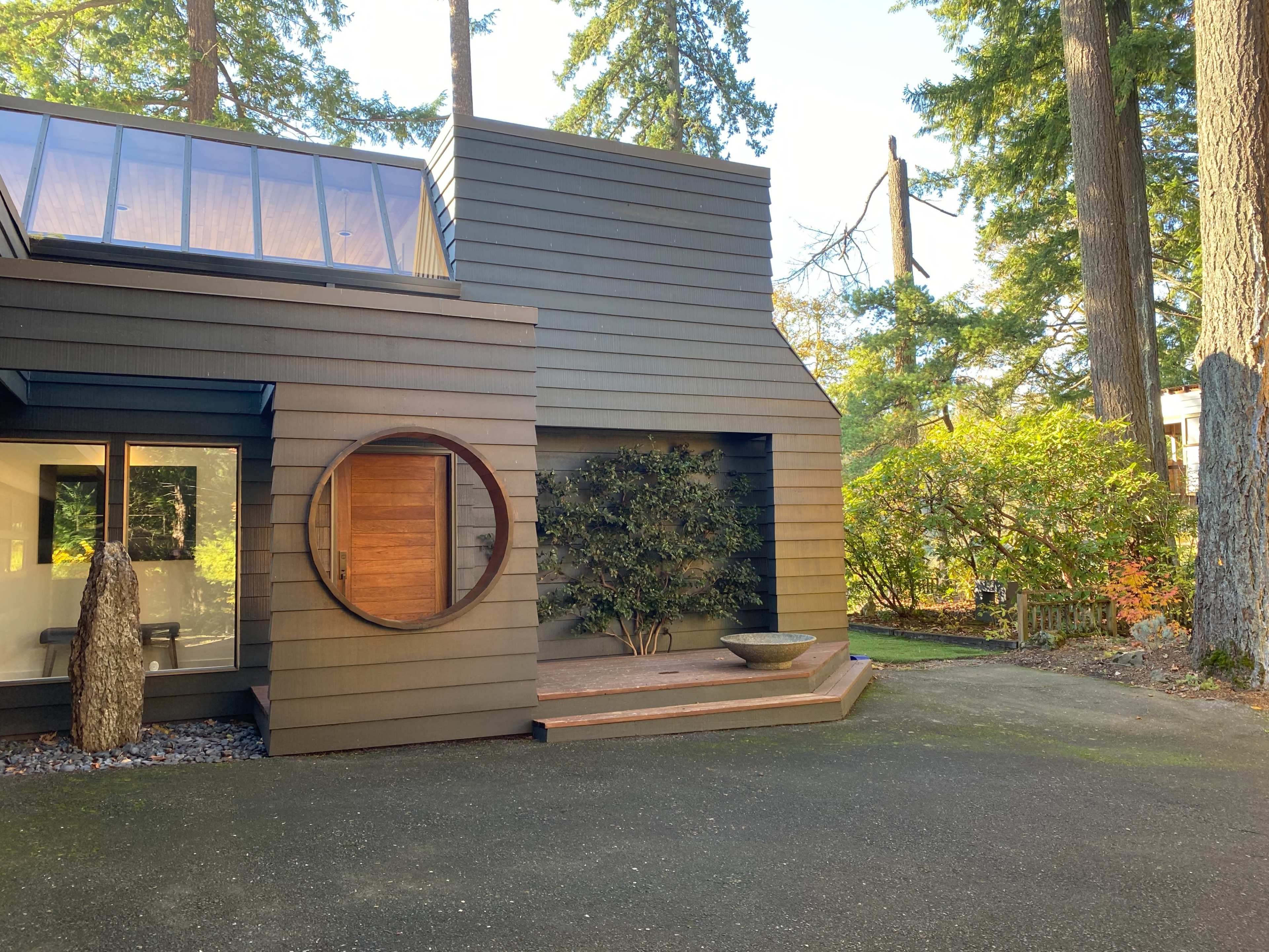 A modern two-story house with wooden siding, featuring a circular window and a small landscaped area in front.