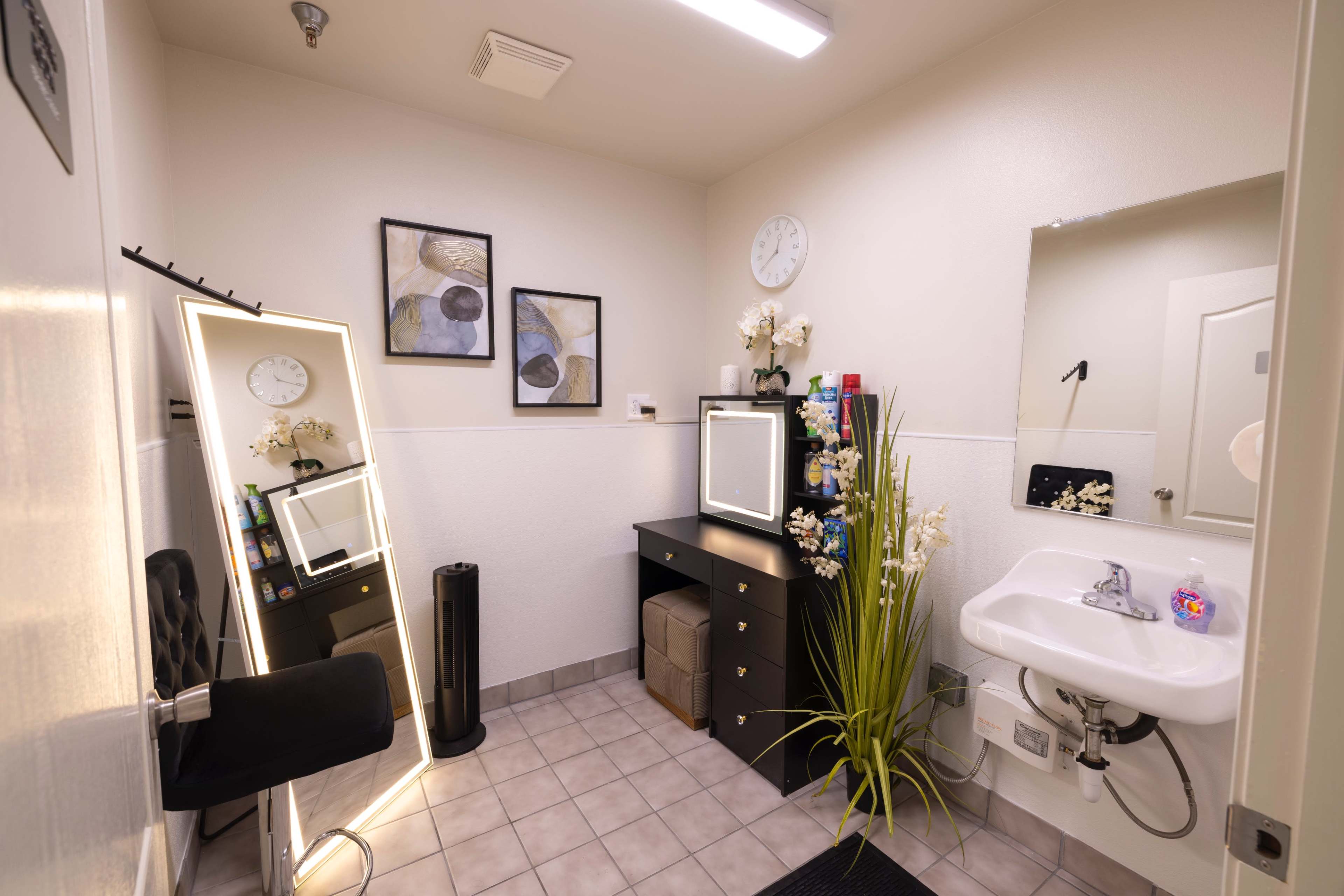 The image shows a clean restroom with a modern sink, a large mirror, a wall clock, and a black storage cabinet containing various personal care items.