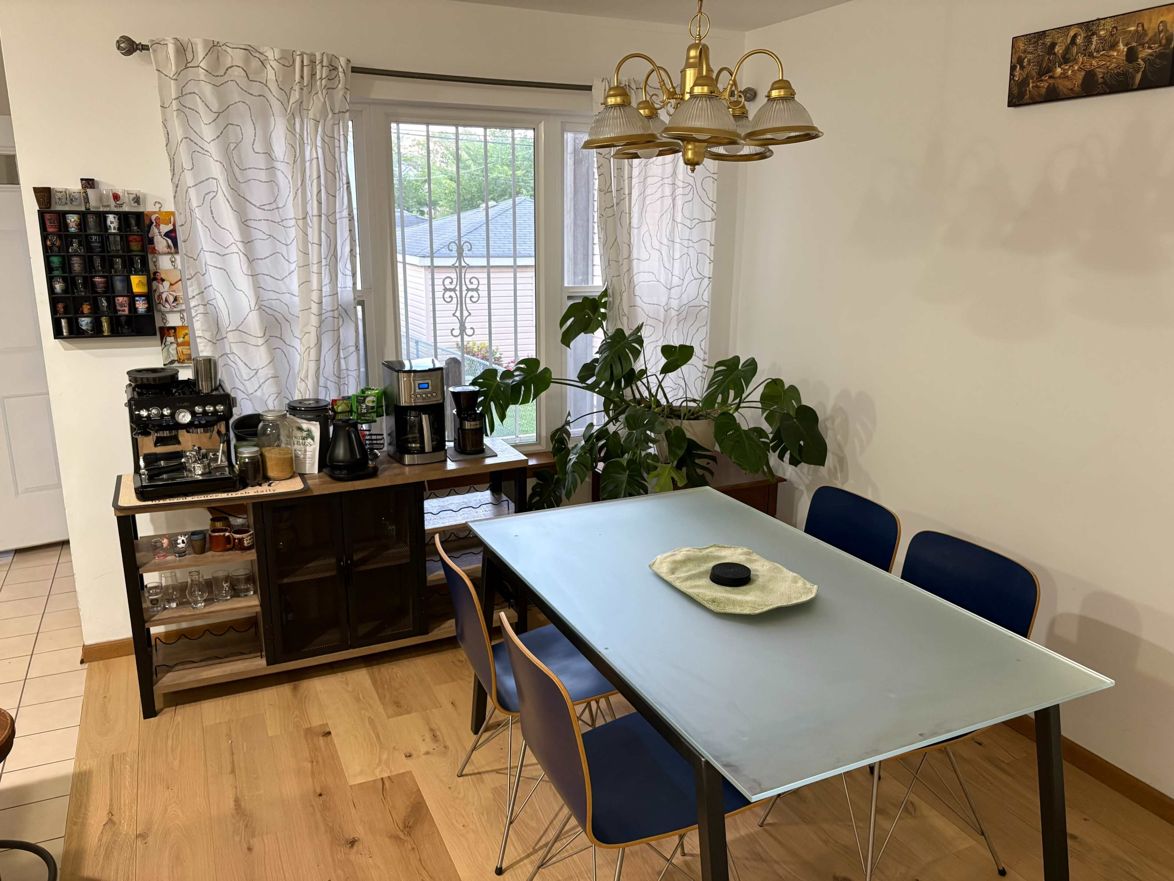 A dining area features a glass table surrounded by chairs, a buffet with a coffee machine, and large windows adorned with curtains.