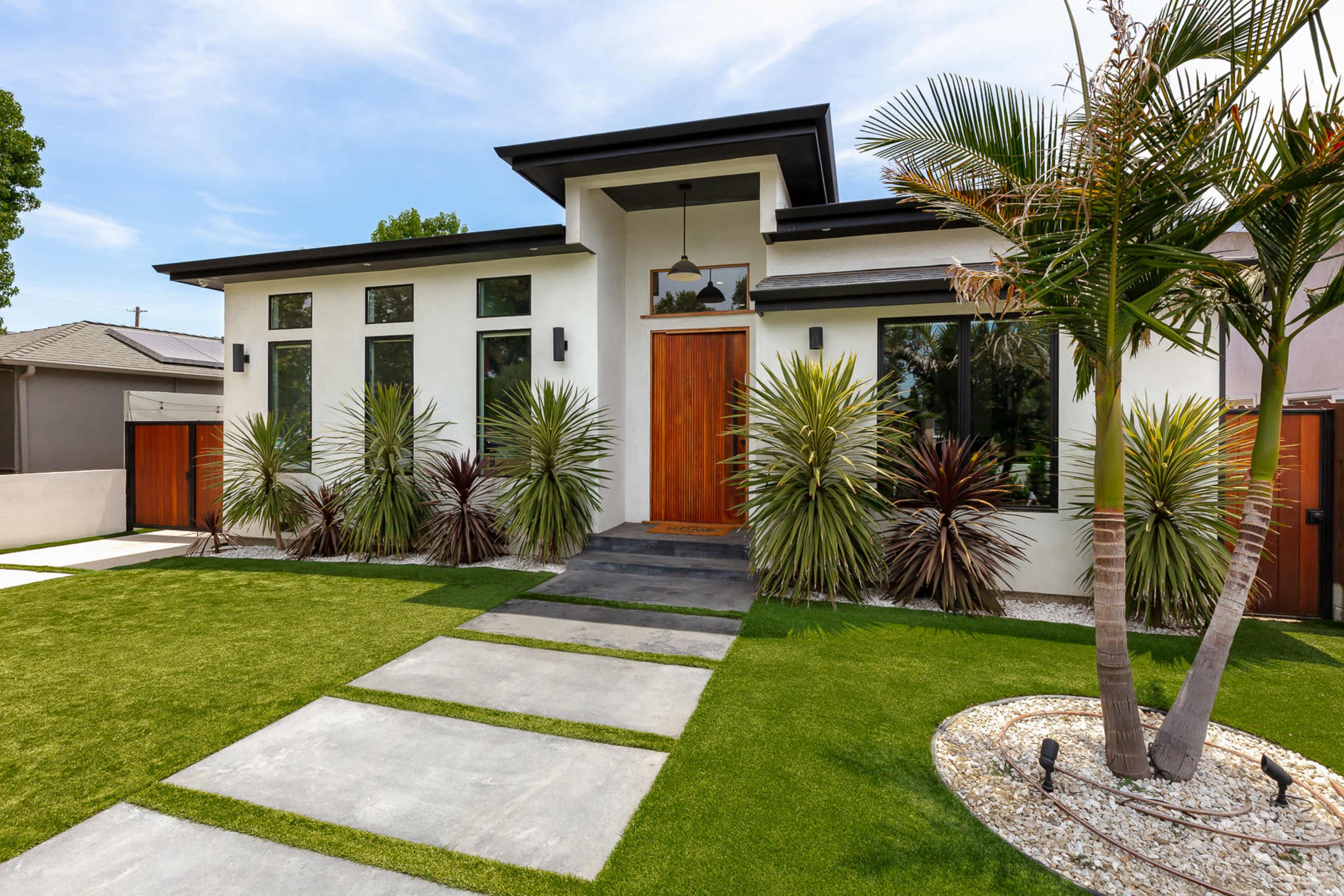 The image shows a contemporary house featuring a wooden front door, large windows, and a landscaped yard with a walkway and various plants.