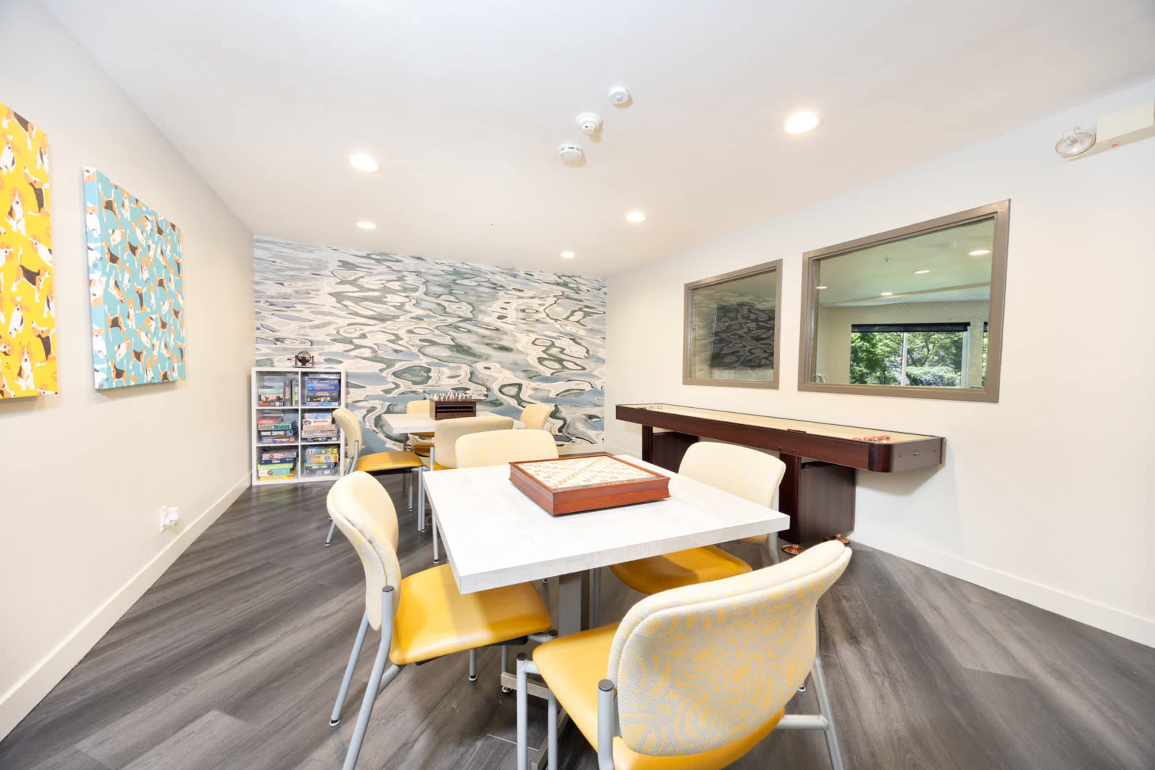 The image shows a modern room with a table surrounded by yellow chairs, a decorative wall mural, and a wooden counter against one wall.