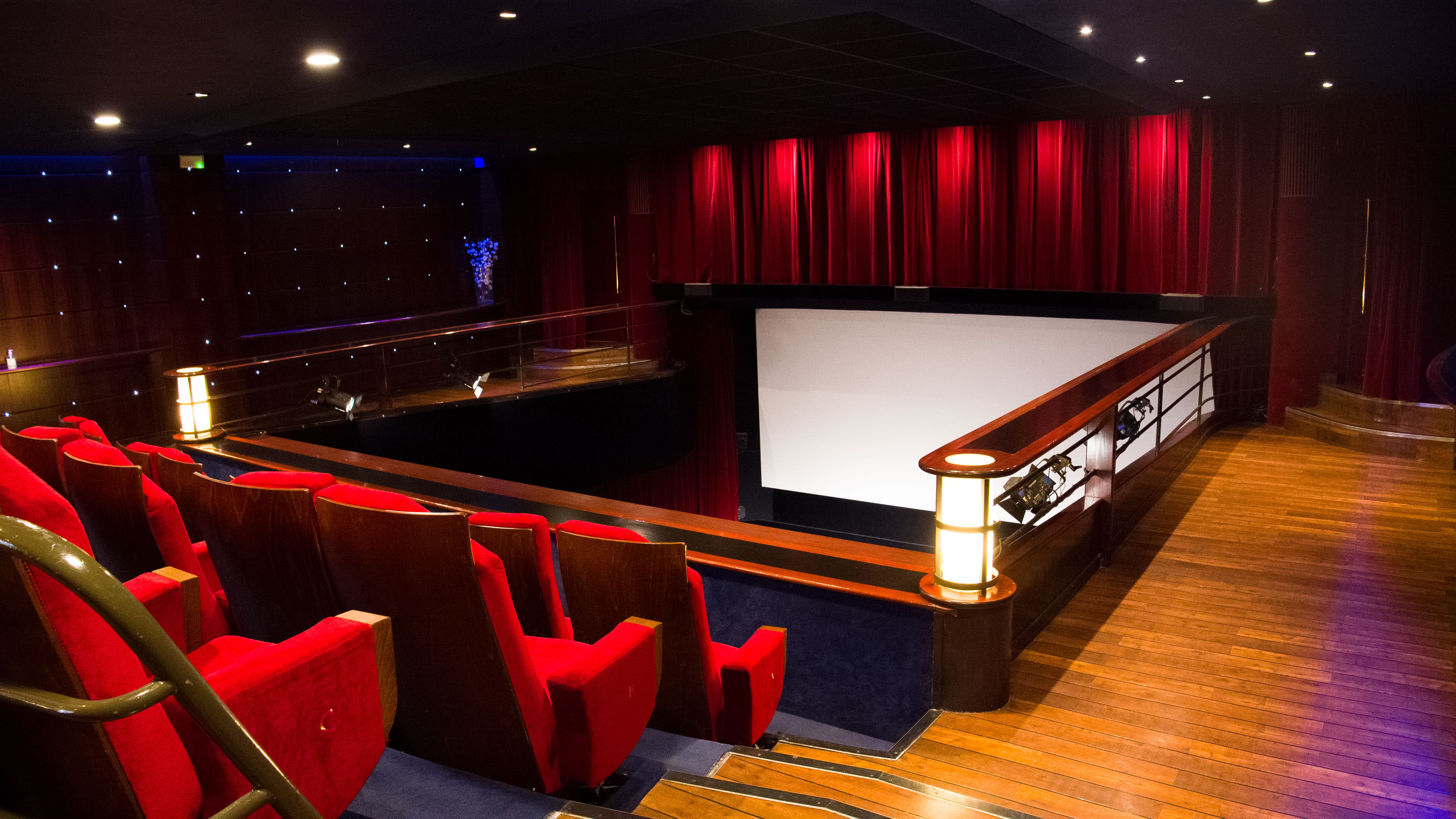 A theater interior with red seating, wooden flooring, and a large blank screen at the front.
