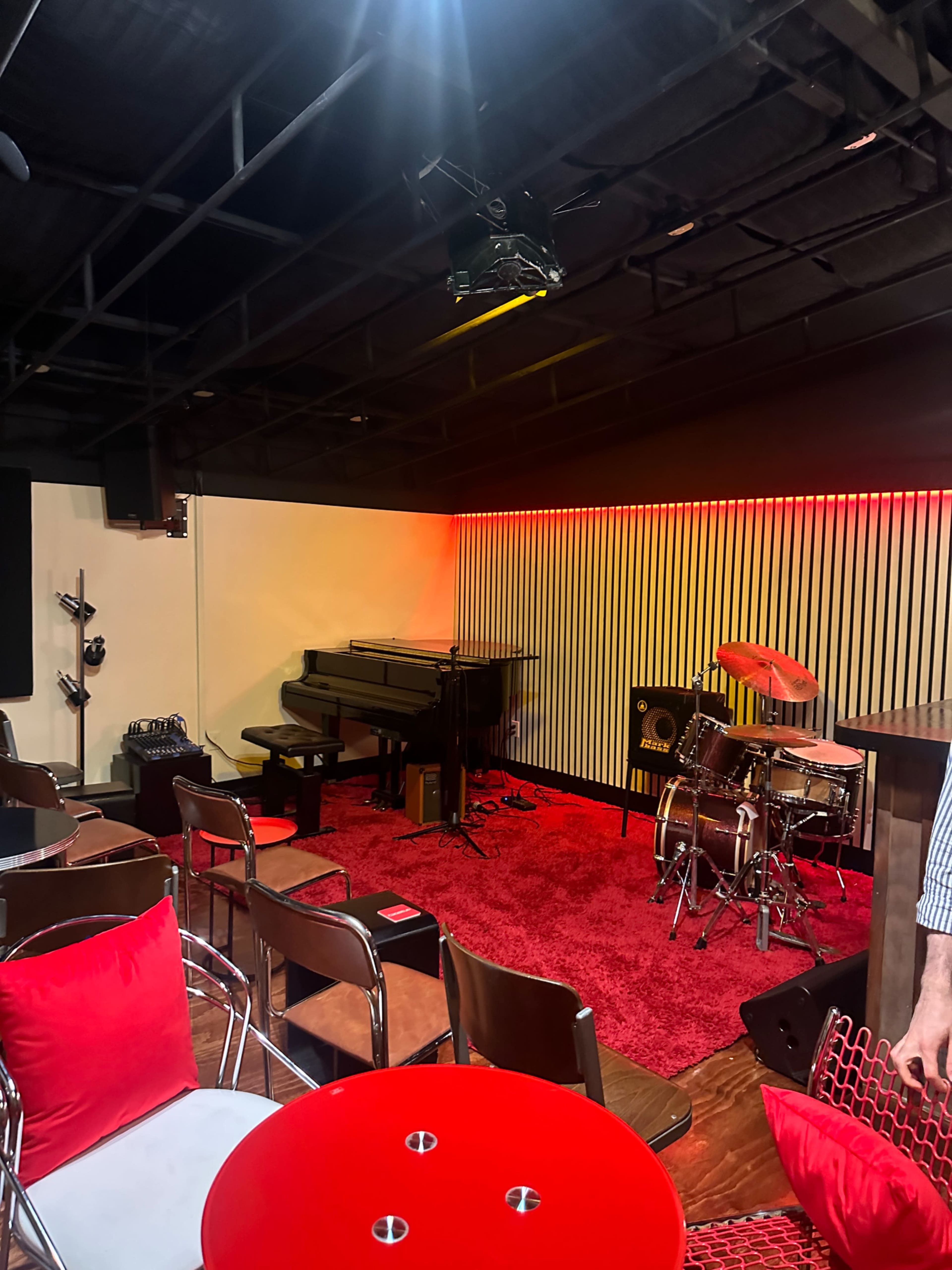 The image shows a dimly lit music venue featuring a piano, a drum set, and seating arrangements with red cushions.