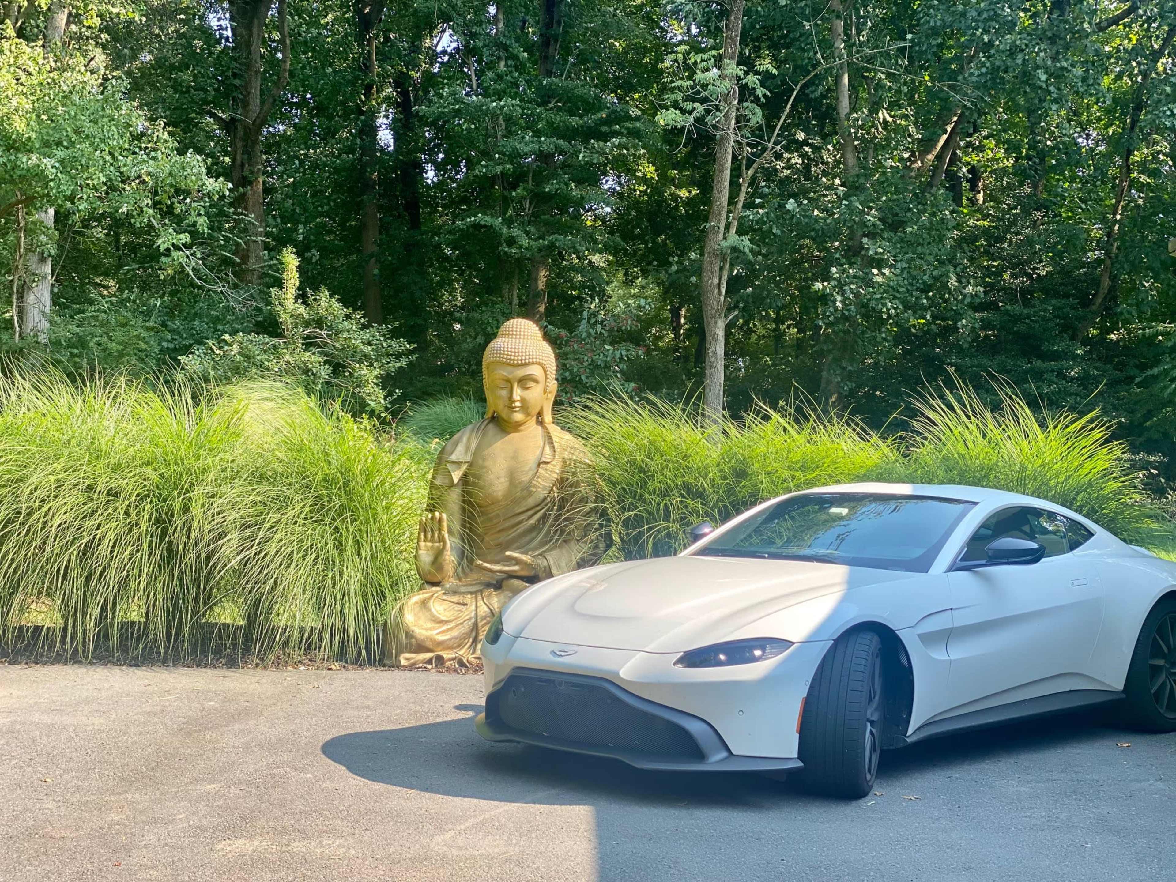 A white sports car is parked beside a large golden Buddha statue set against lush greenery.