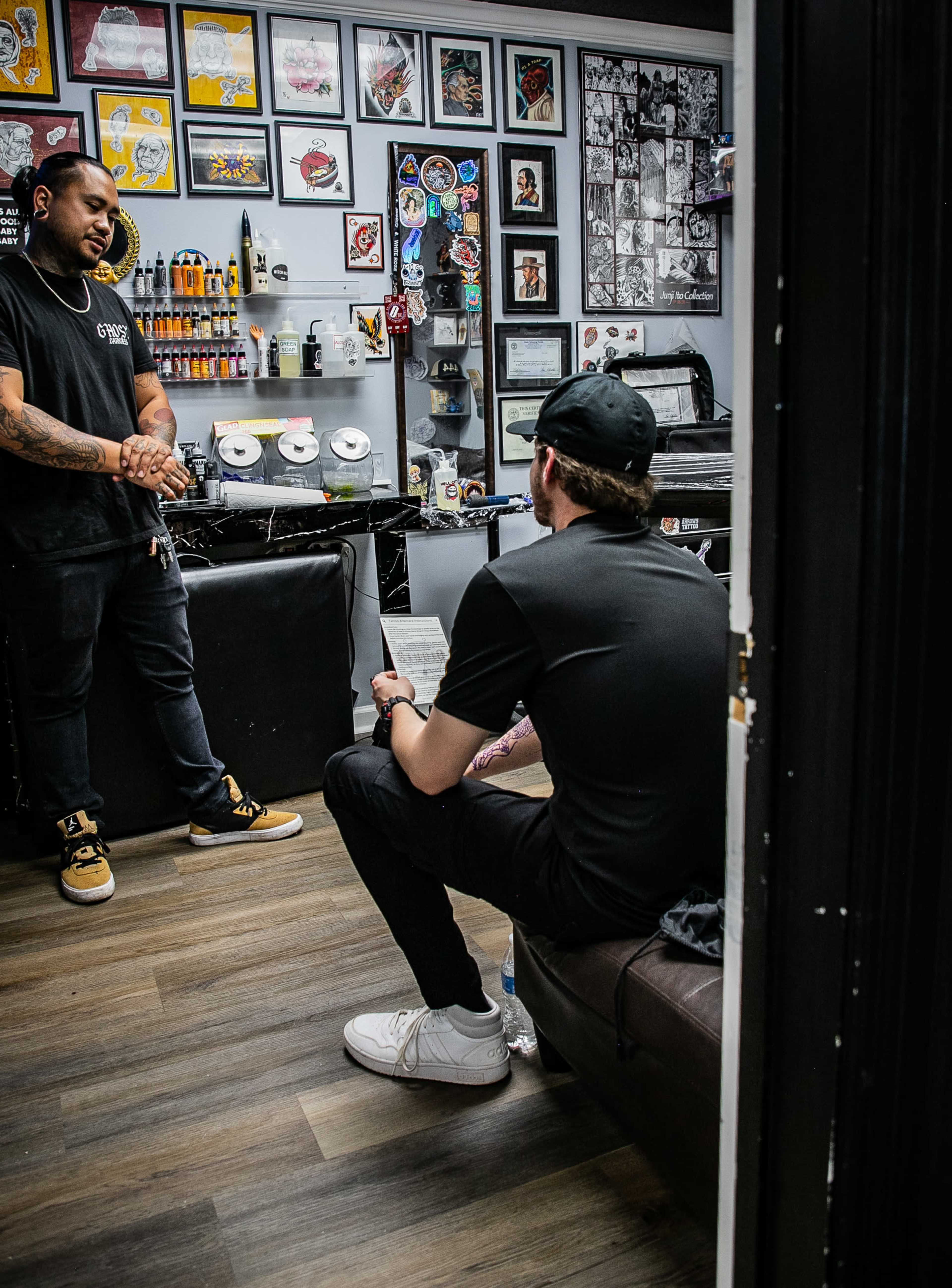 A tattoo artist stands beside a work station while a client sits on a couch, reviewing paperwork in the studio.
