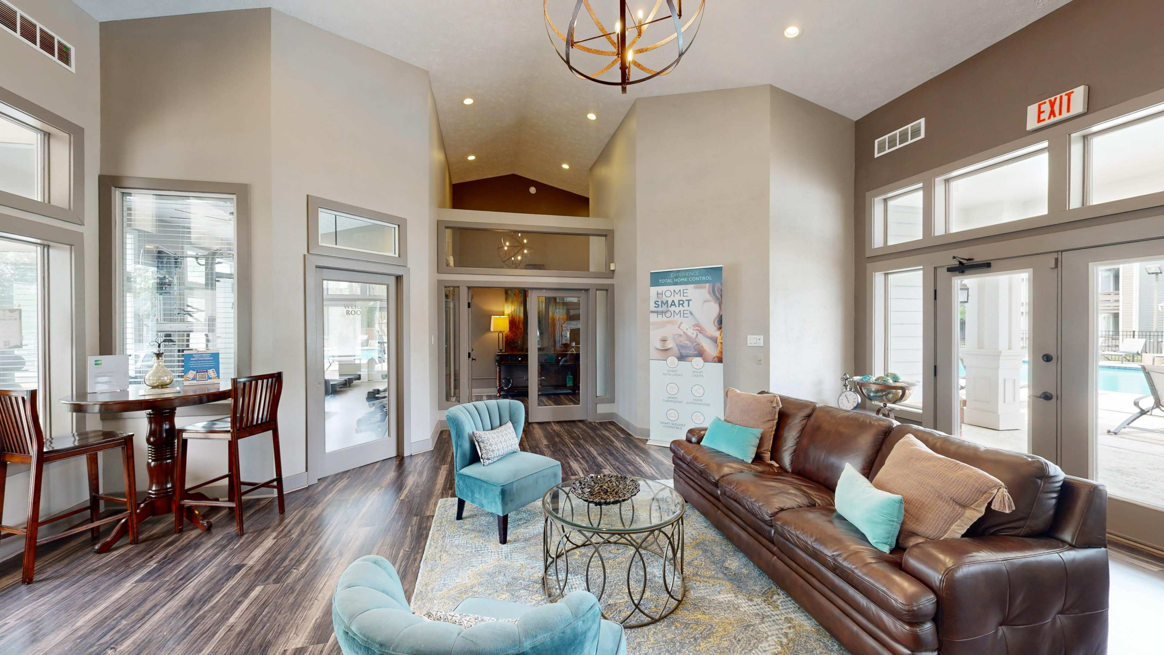 The image shows a modern lobby with a brown leather sofa, light blue chairs, and a decorative rug, featuring large windows and a high ceiling.