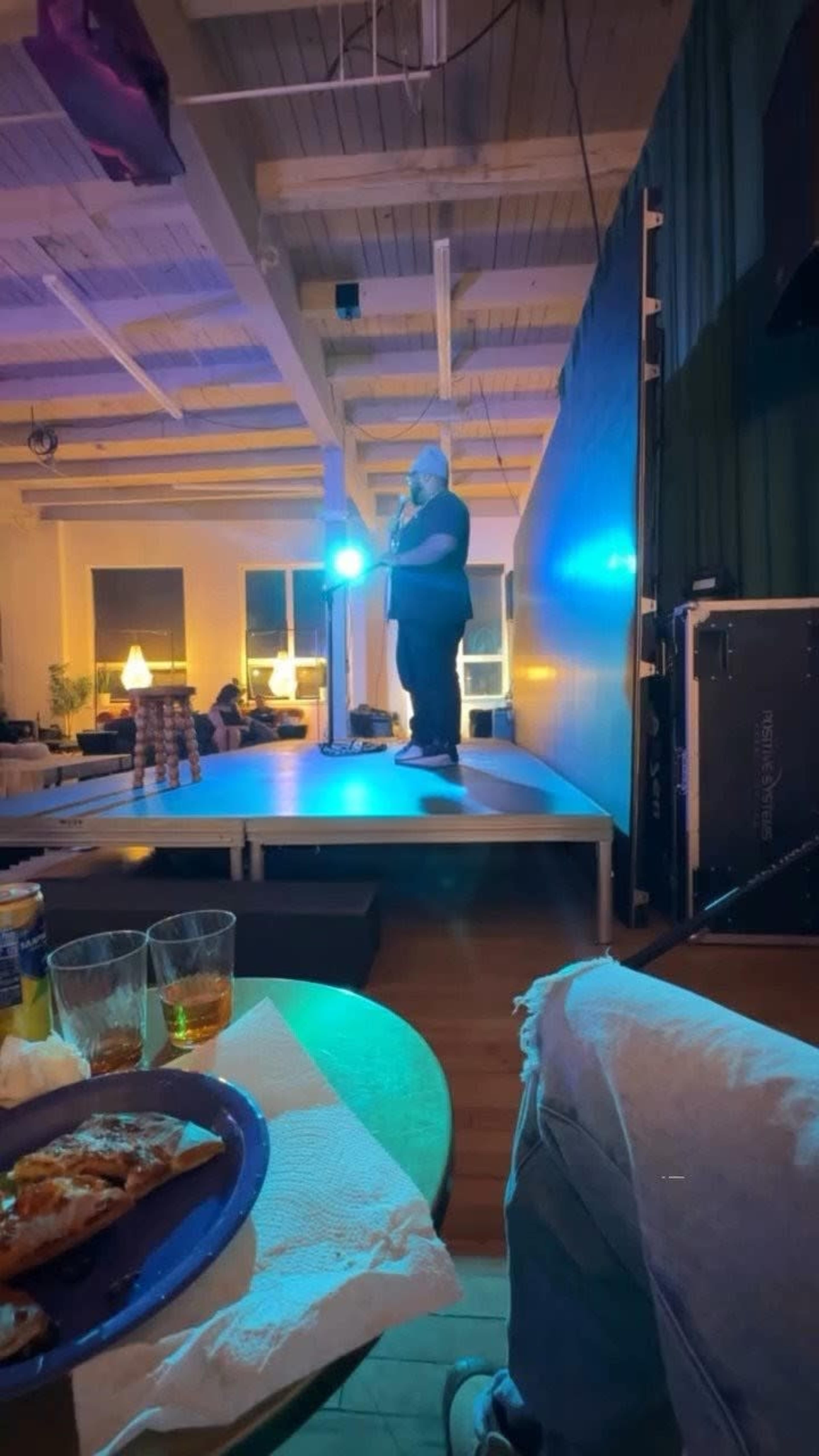 A performer stands on a stage with a microphone under a spotlight, while an audience sits at tables in a well-lit venue.