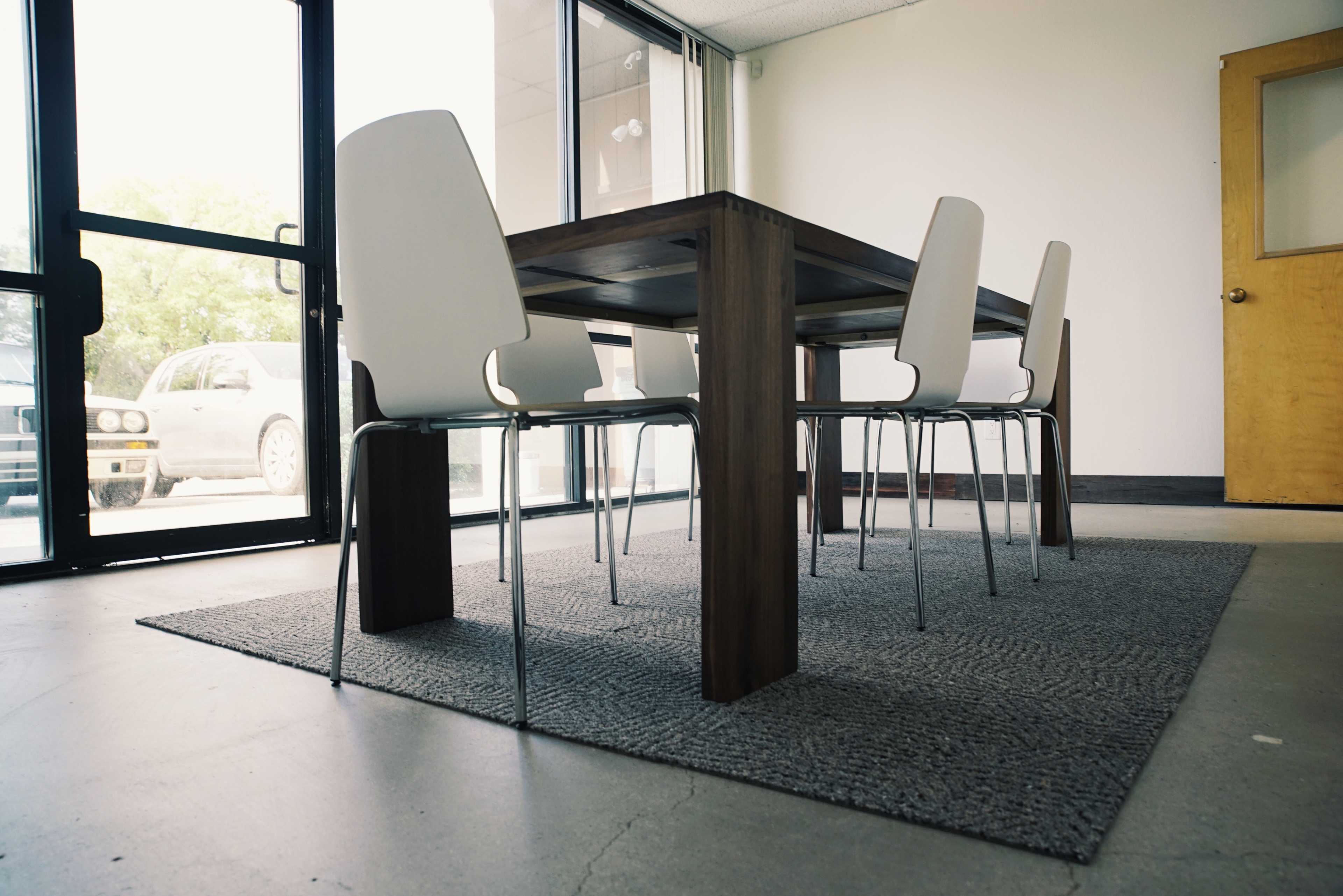 A wooden table with four metal chairs sits on a gray rug in a spacious, well-lit room with large windows.