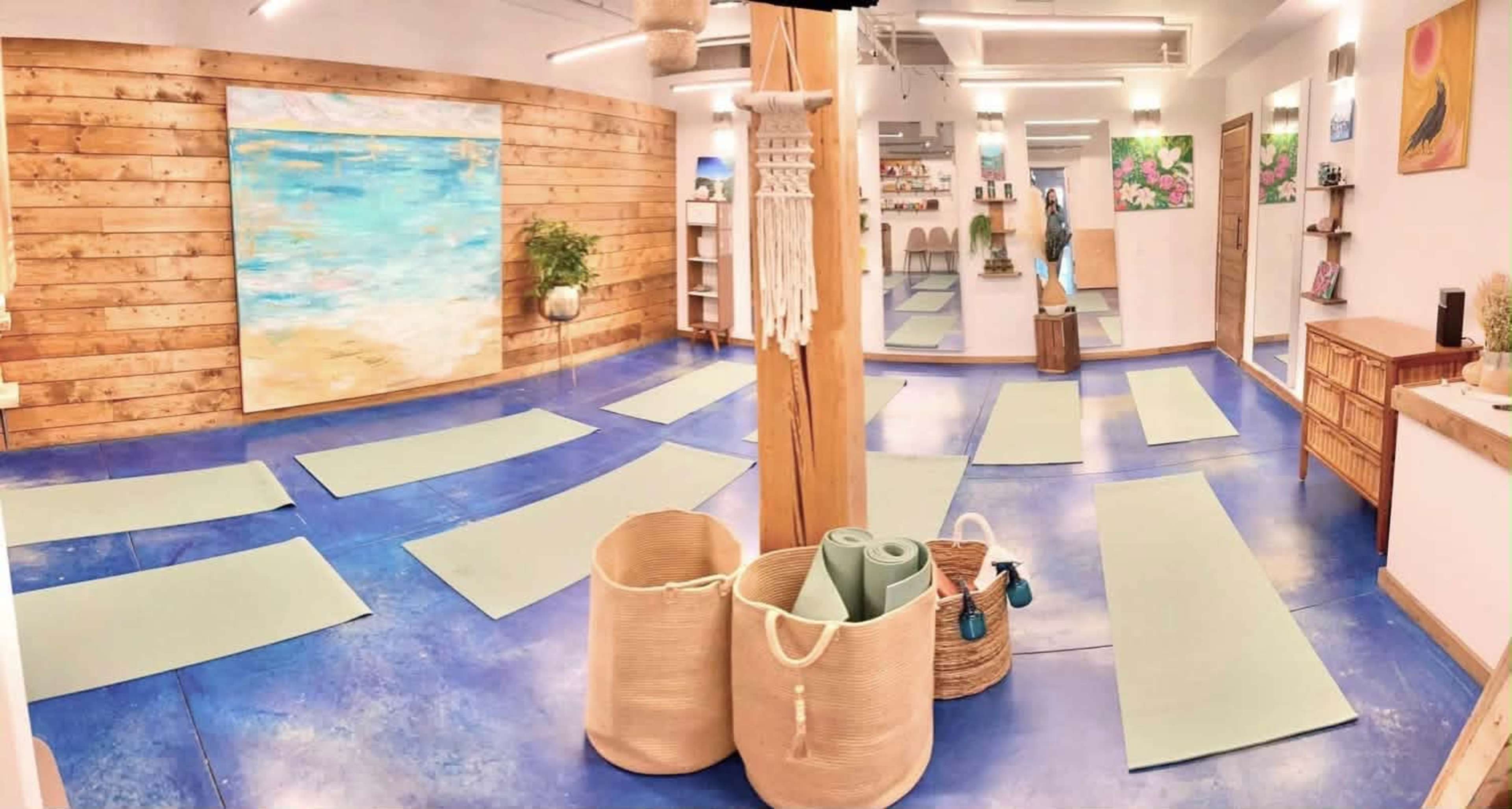 The image shows a yoga studio with several yoga mats arranged on a blue floor, wooden walls, and decorative plants, accompanied by artwork on the walls.