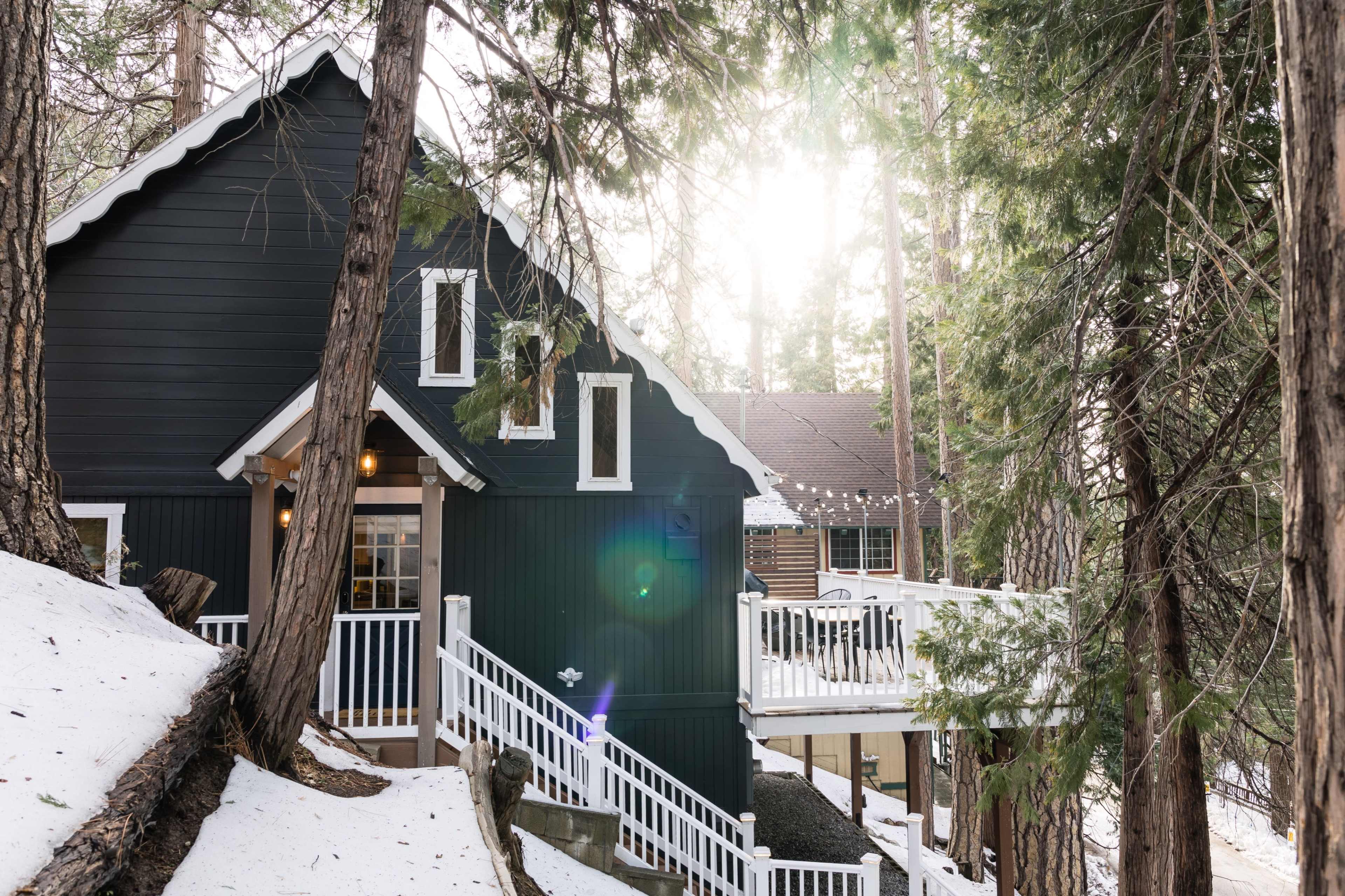 A dark-colored cabin with white trim is nestled among tall trees, with snow covering the ground and sunlight streaming through the branches.