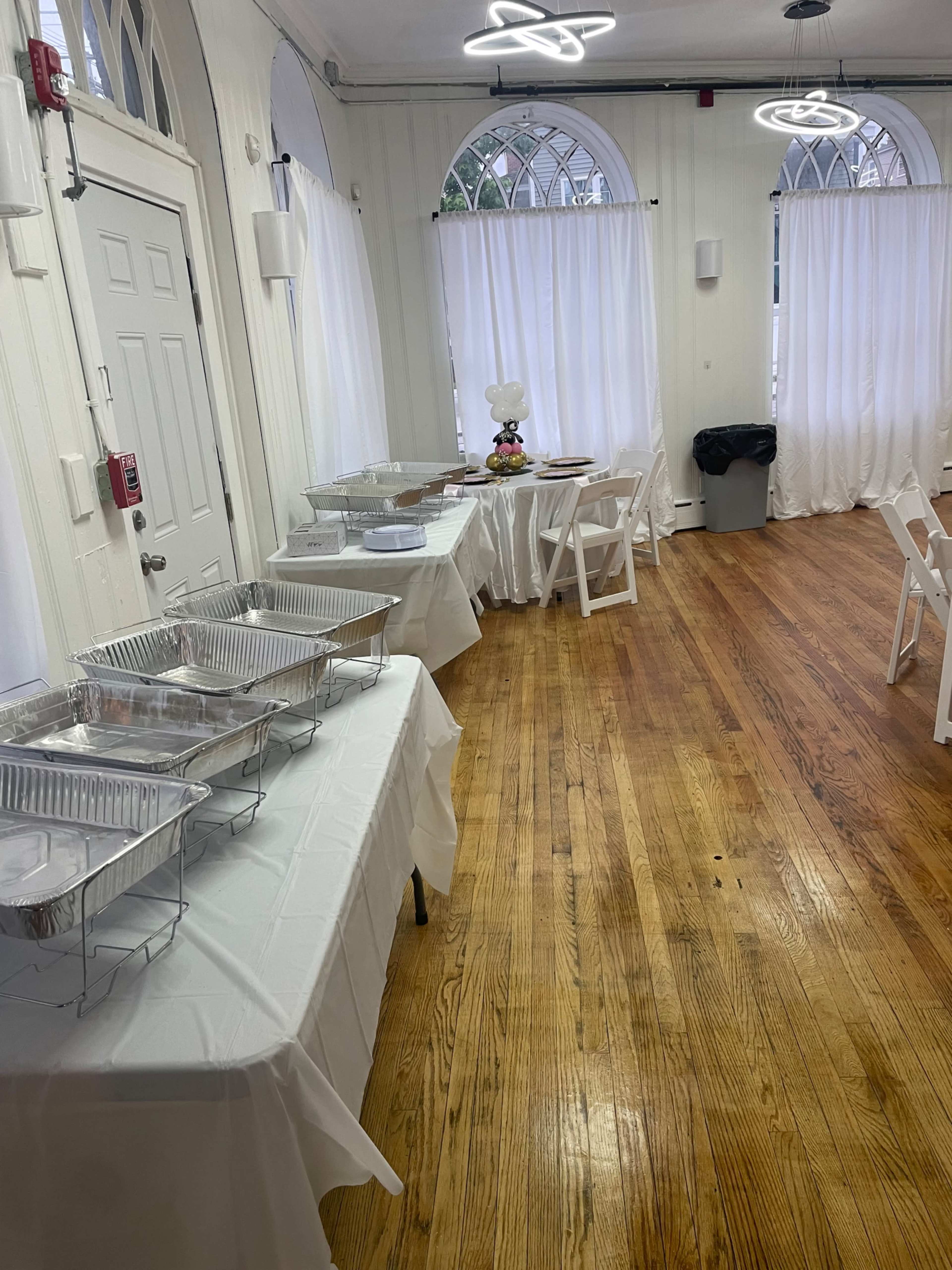 An indoor event space with tables covered in white cloths, arranged with metal food containers, and chairs set up alongside large windows draped with white curtains.