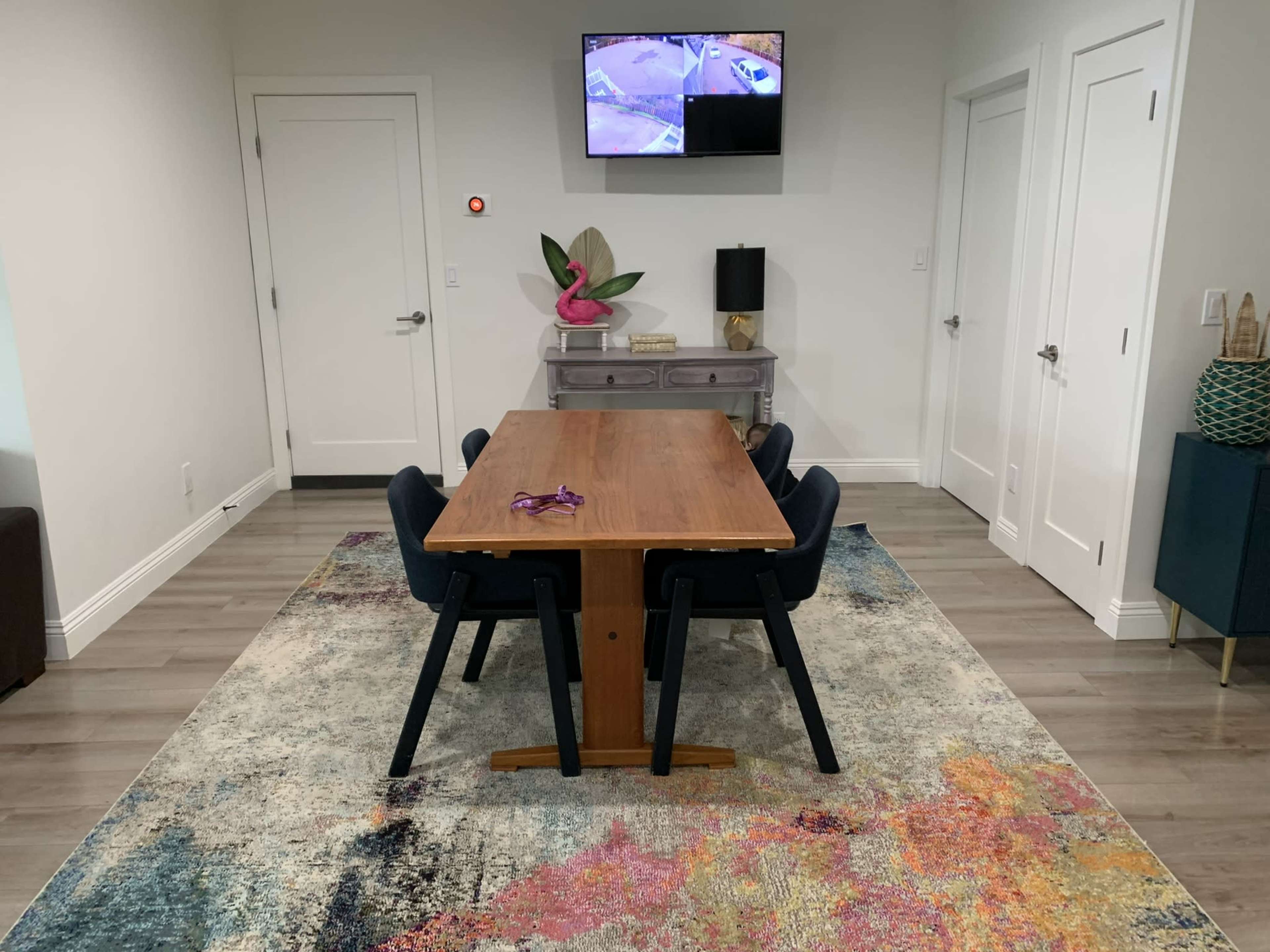 A dining table with black chairs is positioned on a colorful rug in a room featuring a wall-mounted television and two white doors.