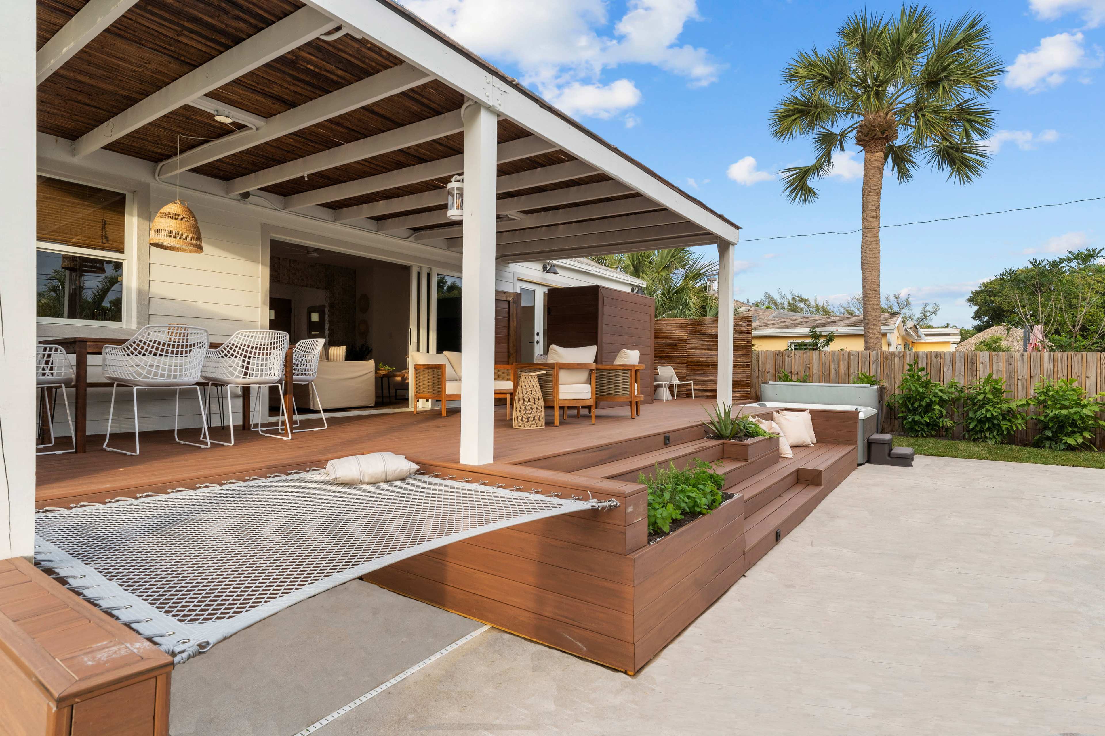 A wooden deck with lounge seating, a dining area, and palm trees surrounds a backyard patio with a hammock and planters.