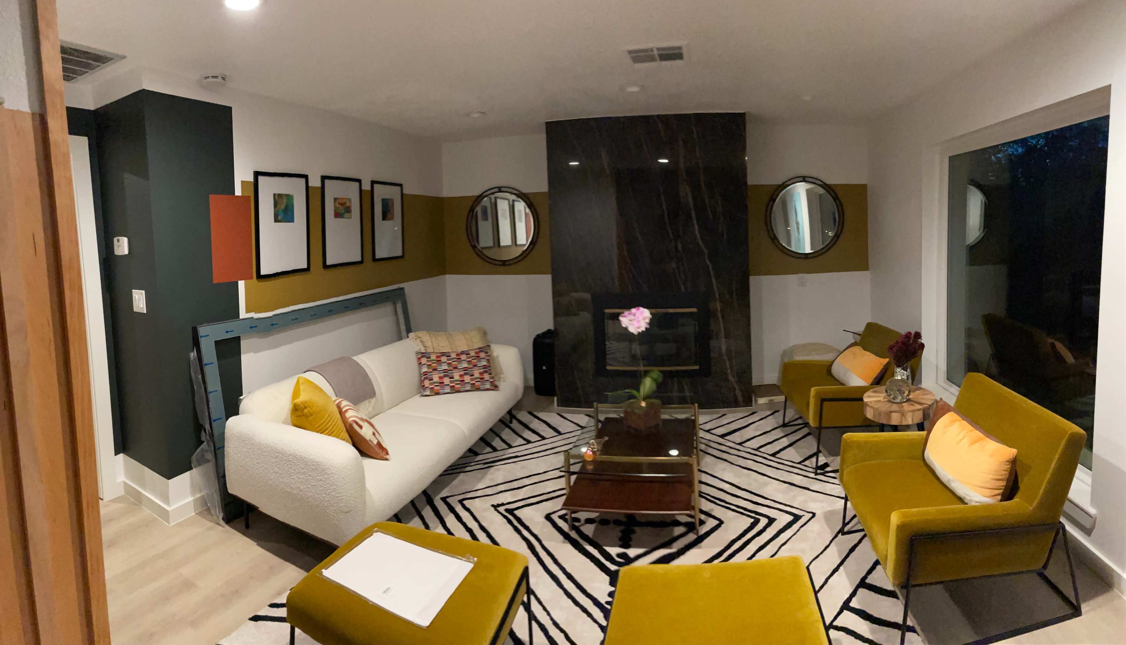 A modern living room features a white sofa, two mustard-yellow chairs, a coffee table with a decorative plant, and framed artwork on the walls, set against a dark stone fireplace.