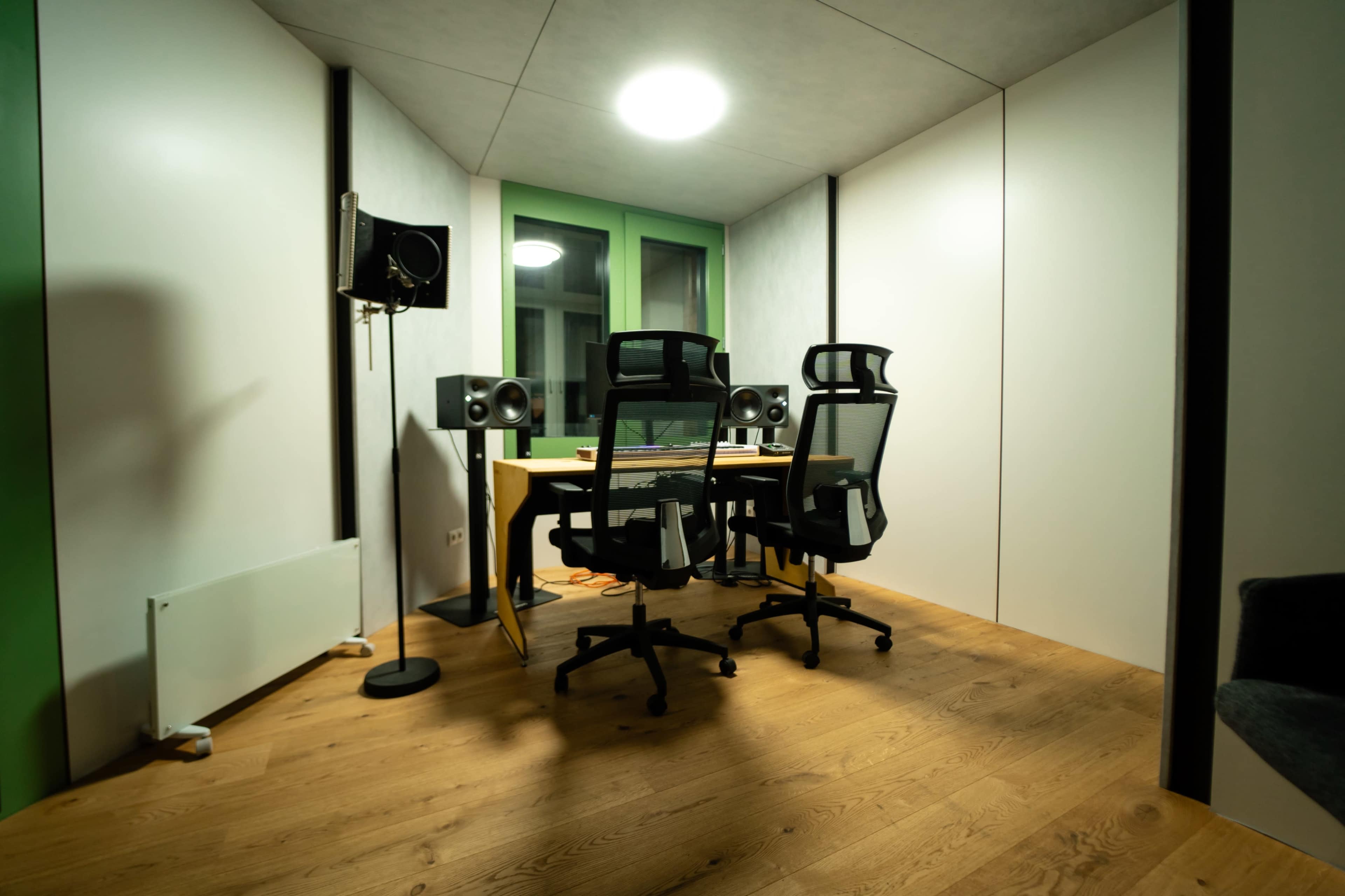 The image shows a modern office space with two task chairs facing a desk equipped with audio speakers and a sound recording setup, enclosed by white walls and wooden flooring.