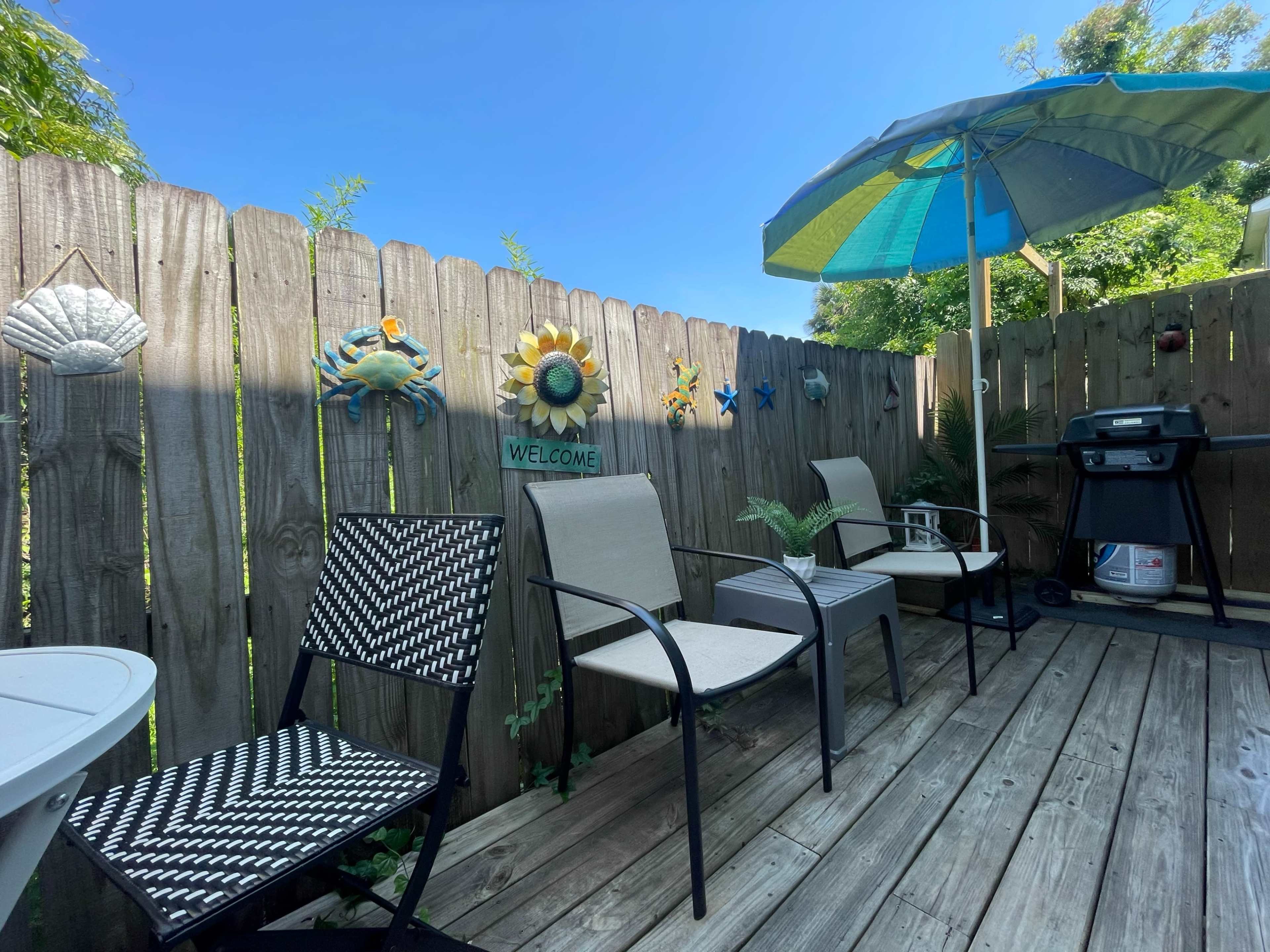The image shows a small outdoor patio area with two chairs, a table, a large umbrella, and a grill, surrounded by a wooden fence adorned with decorative items.