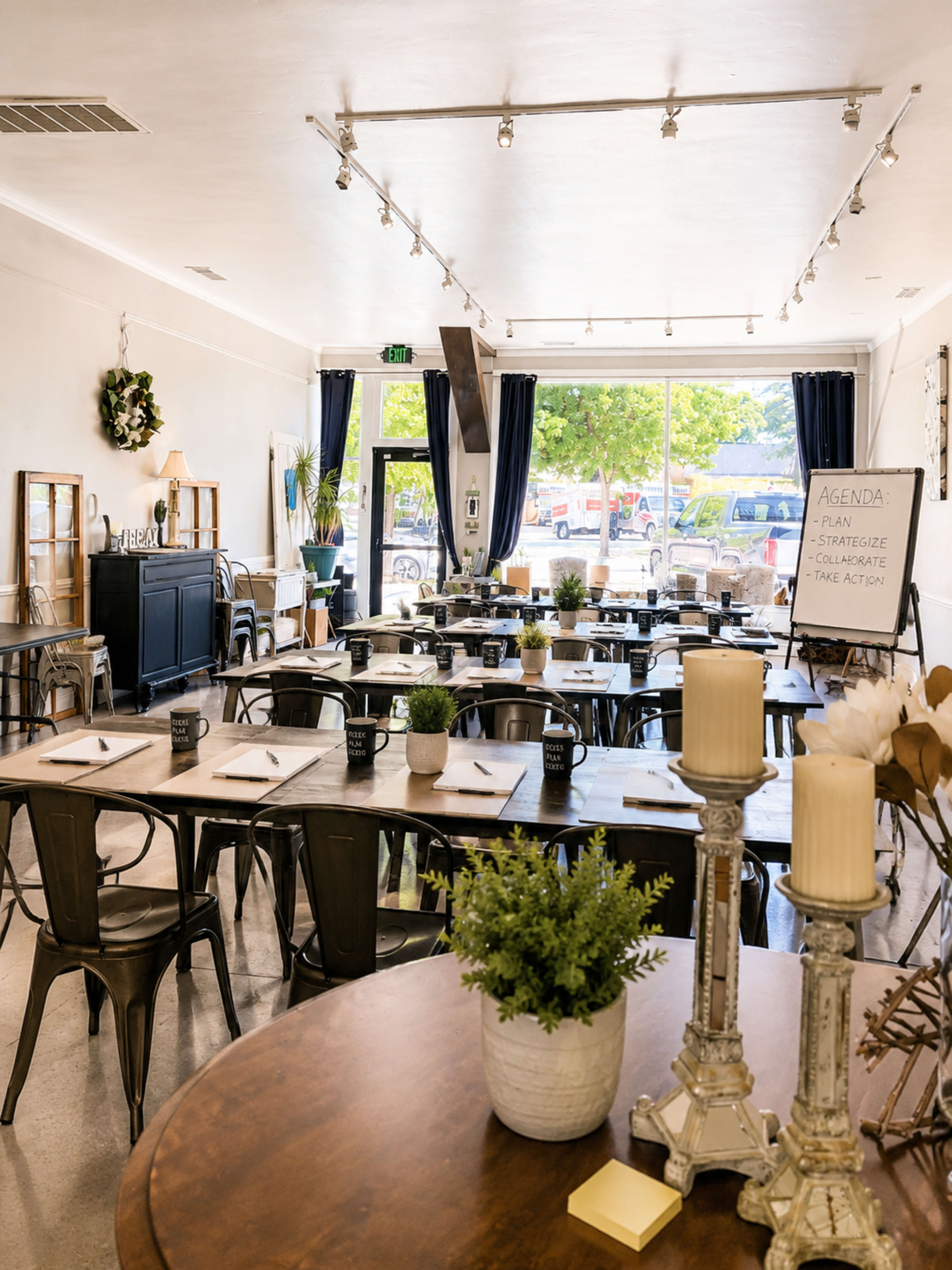 The image shows a spacious room set up for a workshop, featuring multiple tables arranged with notebooks and plants, along with a large whiteboard displaying an agenda.