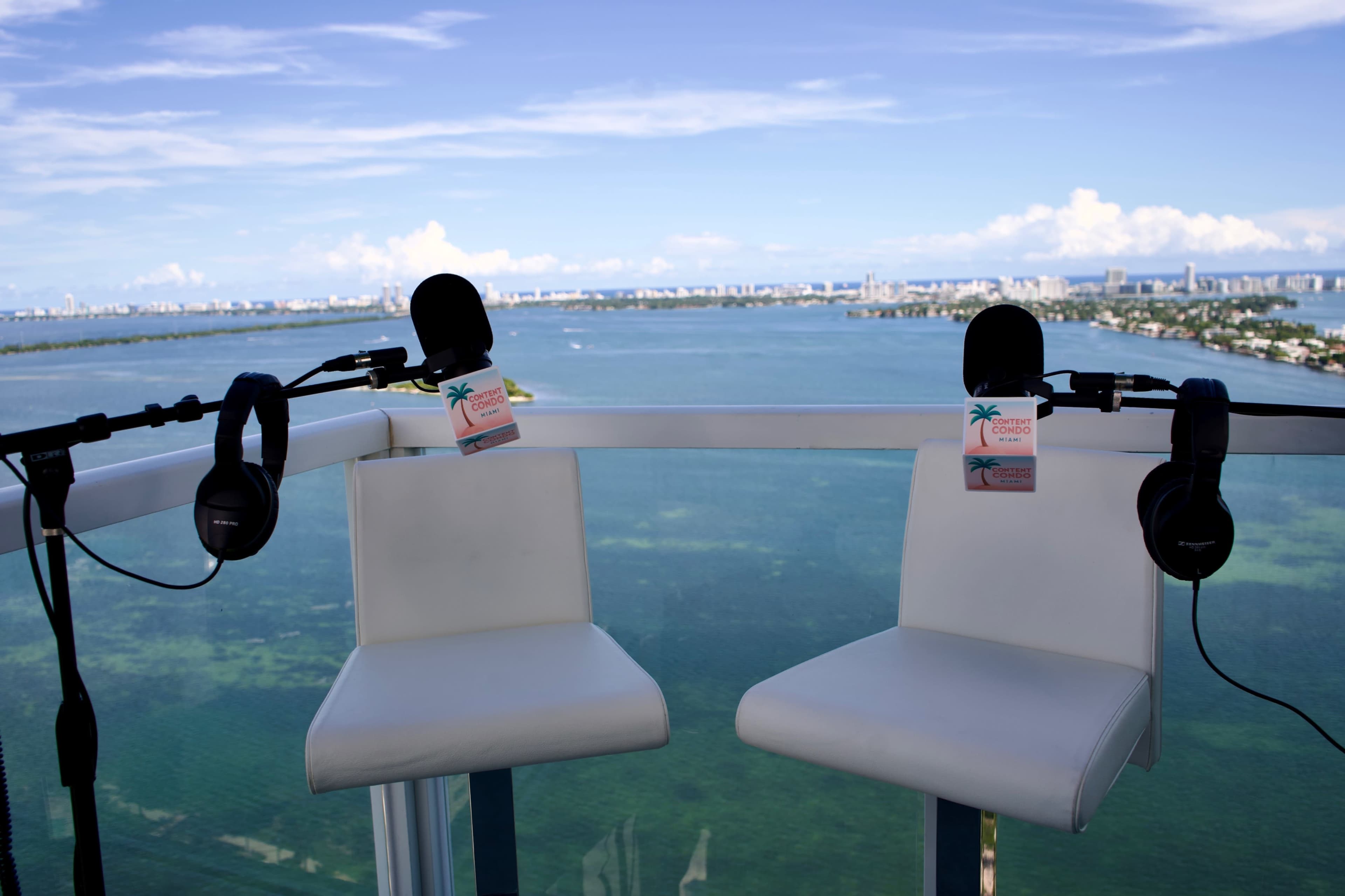 Two white barstools with microphones and headphones are set on a balcony overlooking a coastal cityscape and water.