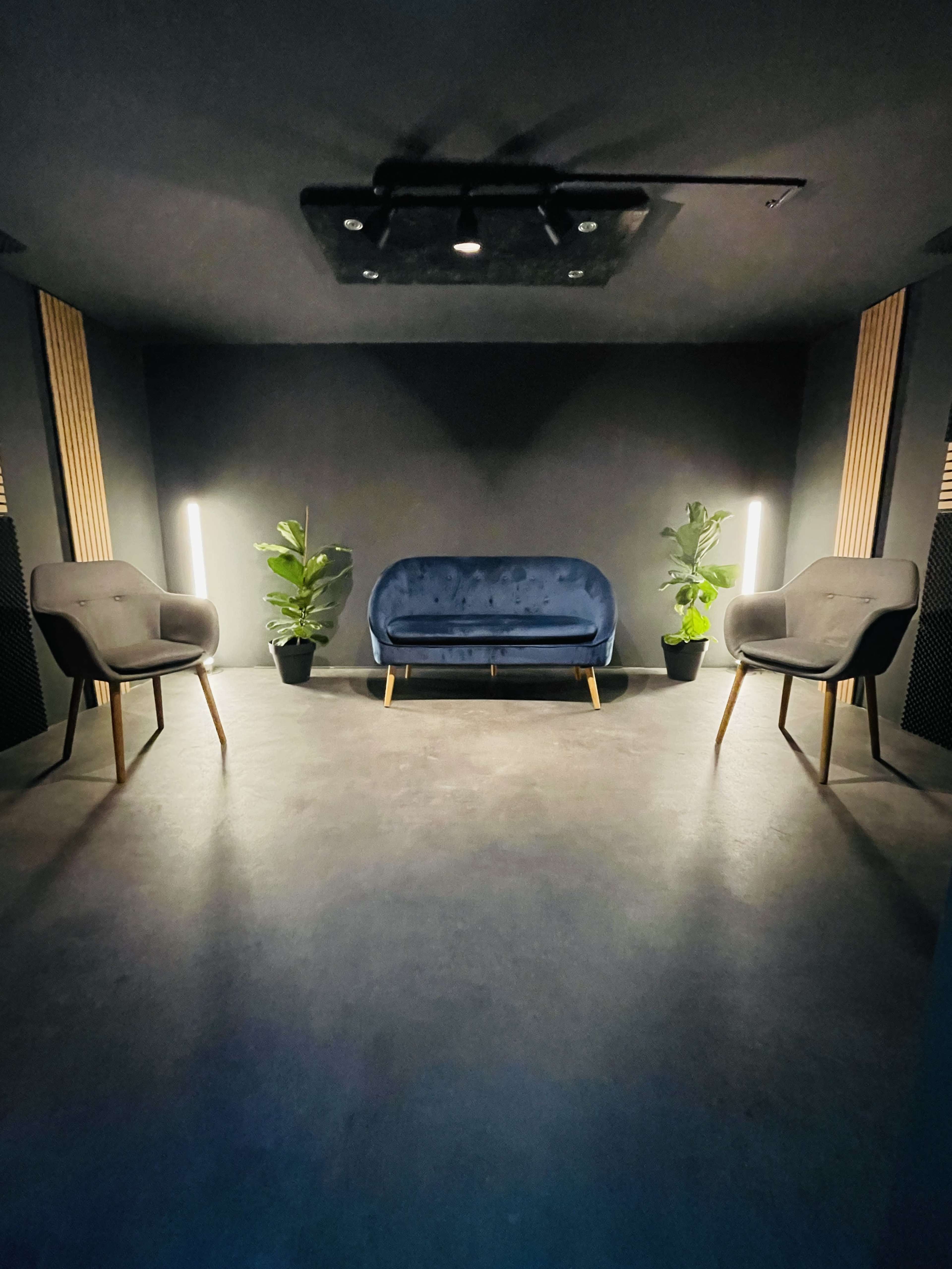 A dark room features a blue sofa and two gray chairs, flanked by potted plants and lit by vertical floor lamps.
