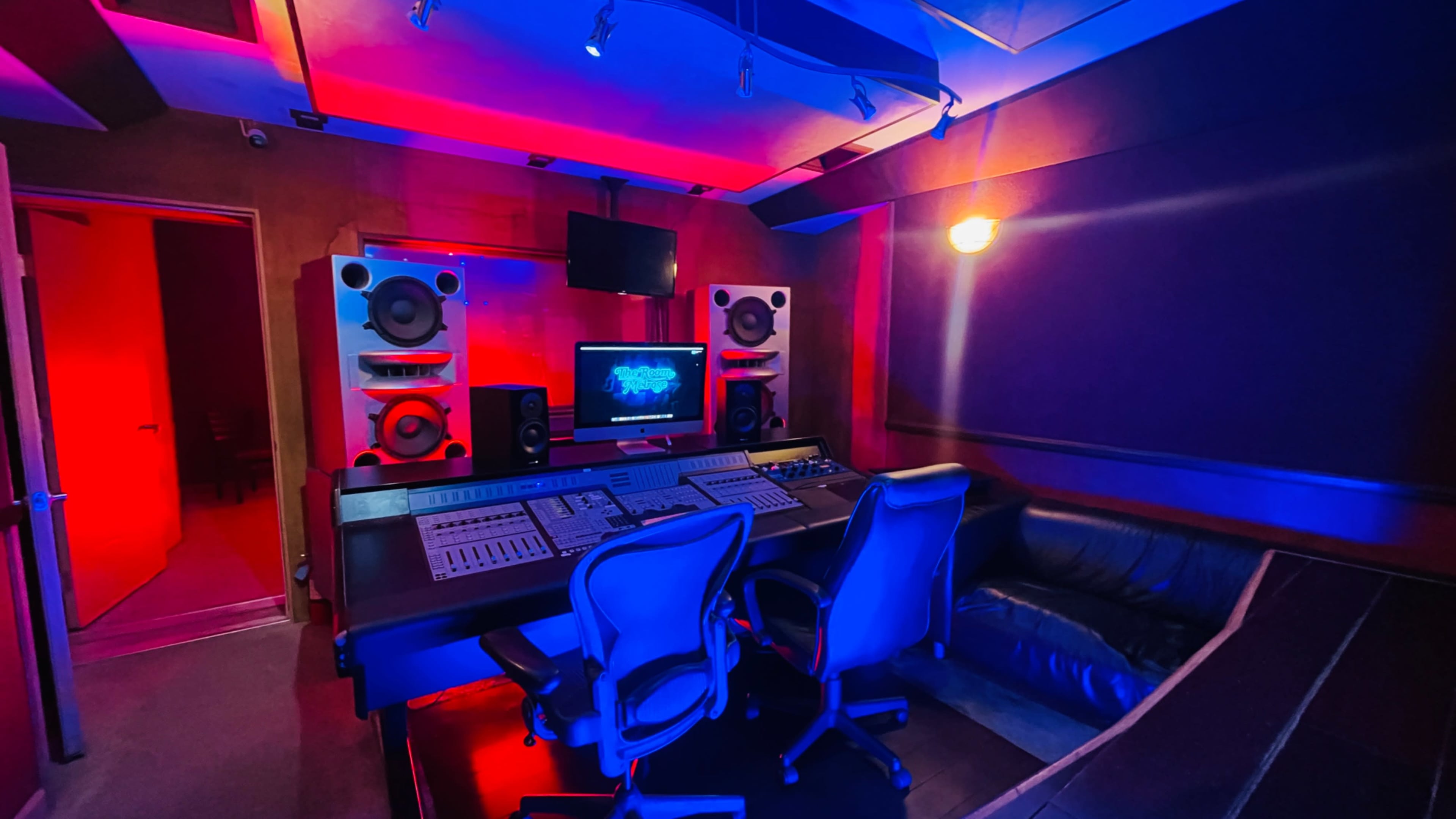 The image shows a music studio with a mixing console, two chairs, large speakers, and colored lighting.