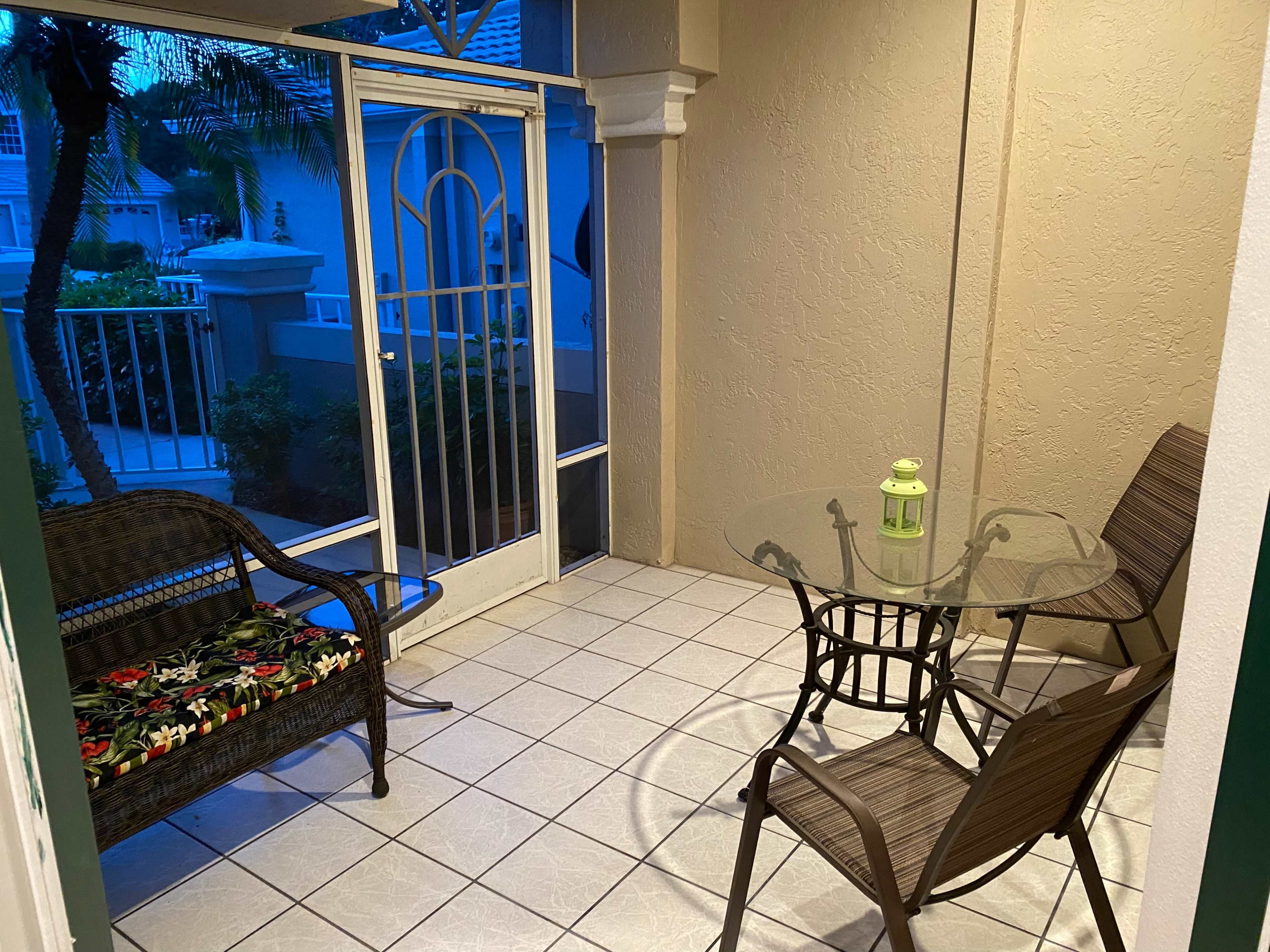 A small enclosed porch features a glass table surrounded by two chairs and a wicker bench, with a lantern on the table and a view of plants outside.