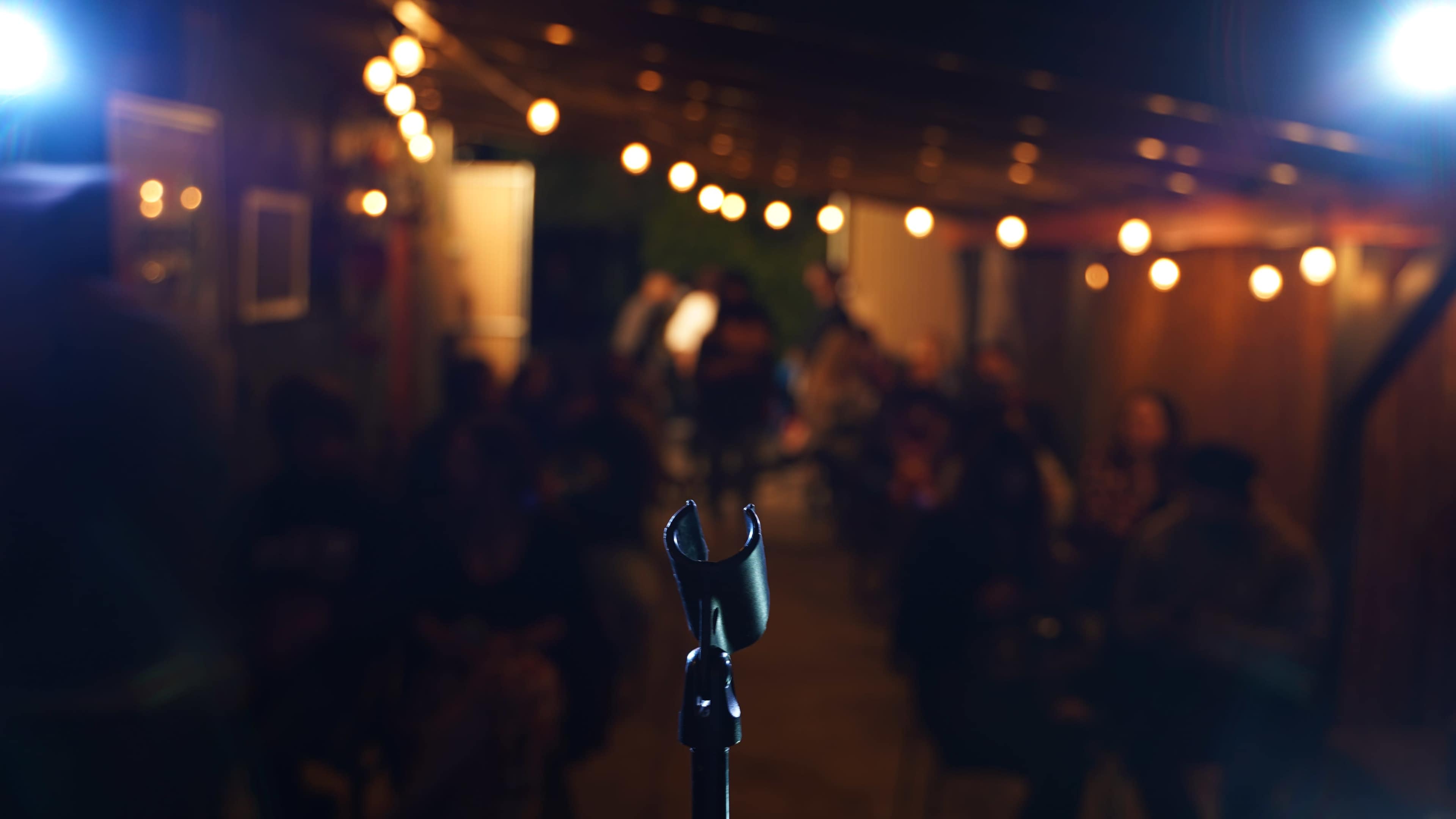 A microphone stands in the foreground, facing an audience seated in a dimly lit space adorned with string lights.