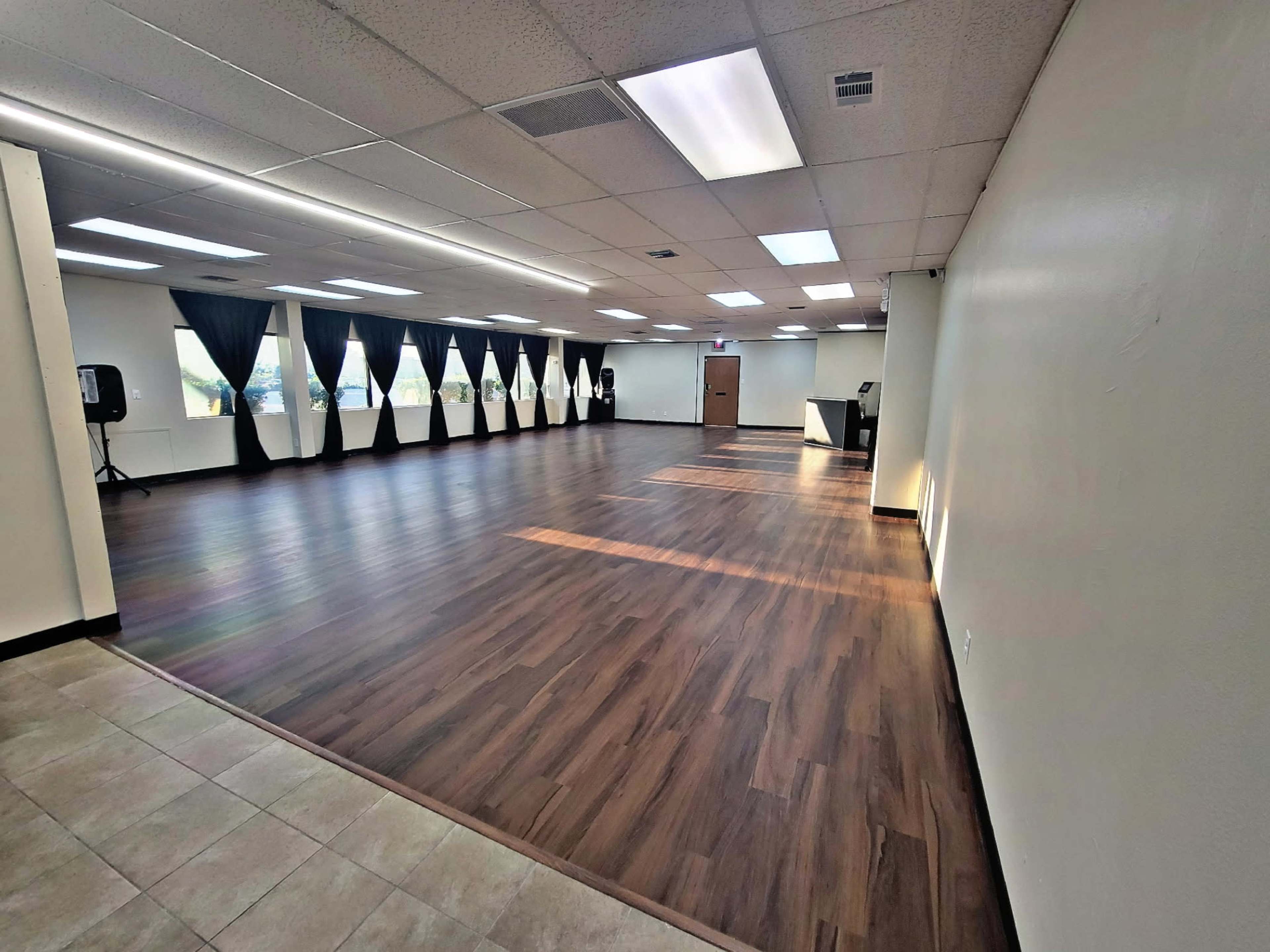The image shows a spacious, well-lit indoor room with wooden flooring and several large windows covered by dark drapes.